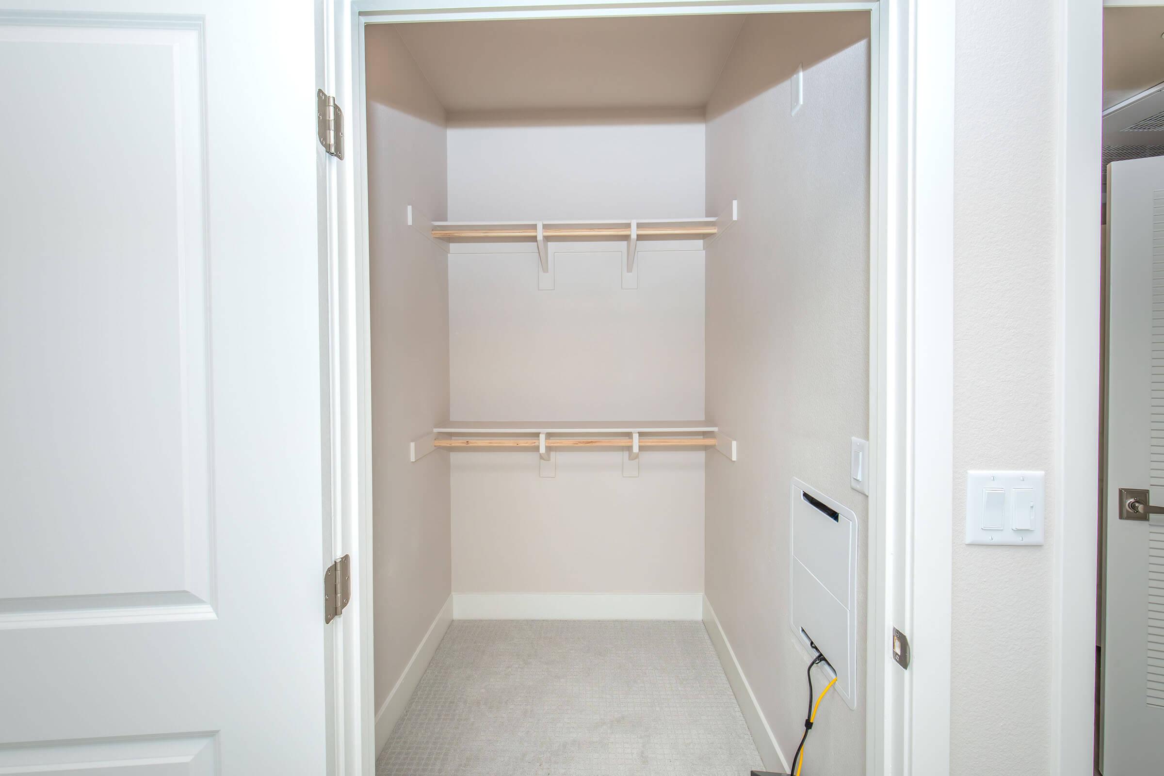 An empty closet with two wooden shelves mounted on the walls. The walls are painted a light color, and the floor is carpeted. A small utility area with wires is visible on the right side. The closet has a white door partially open, revealing the unoccupied space inside.
