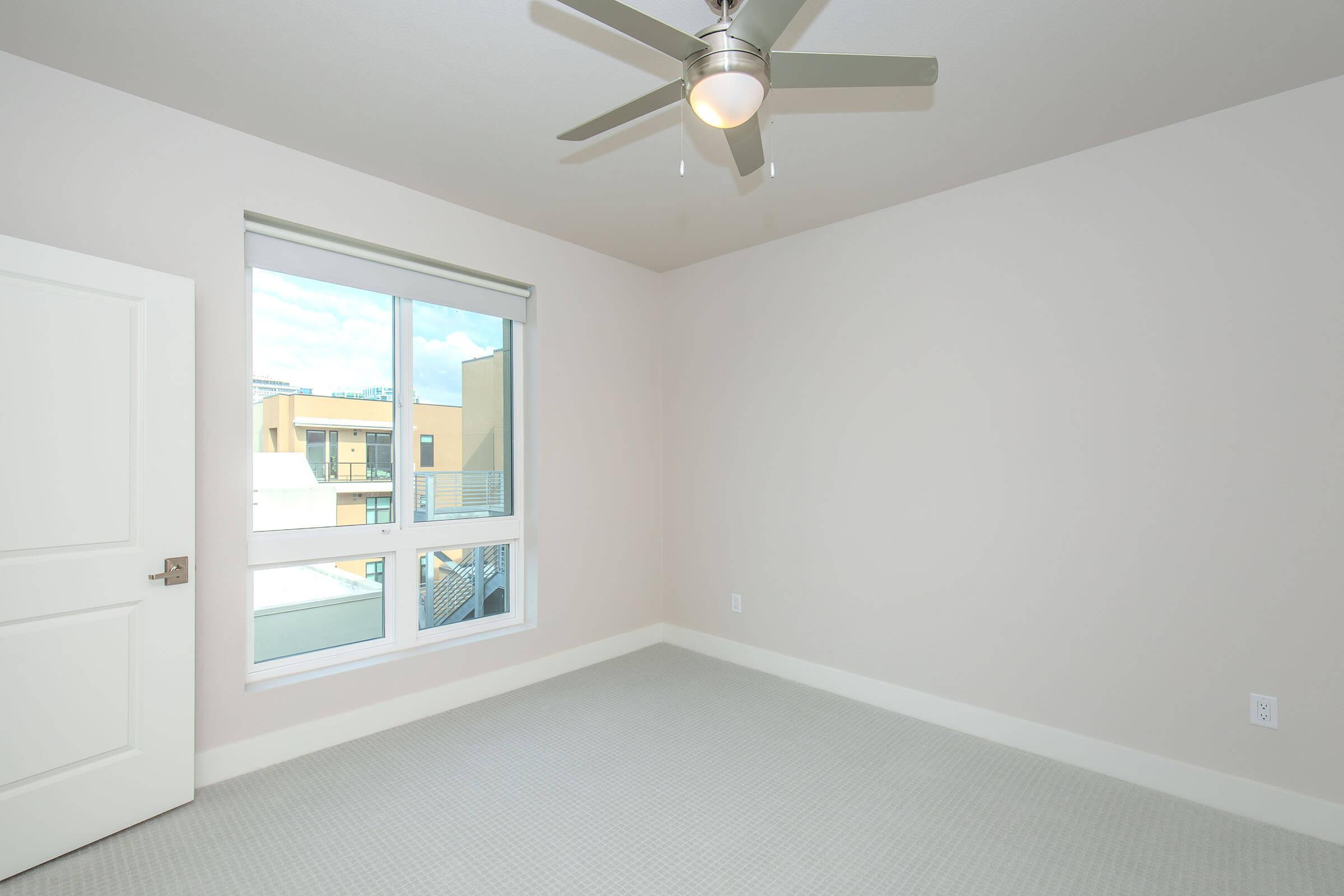 A small, empty bedroom featuring light beige walls, a ceiling fan, and a large window that allows natural light. The floor is covered with a light-colored carpet, and there is a white door on the left side of the room. The overall ambiance is bright and minimalistic.
