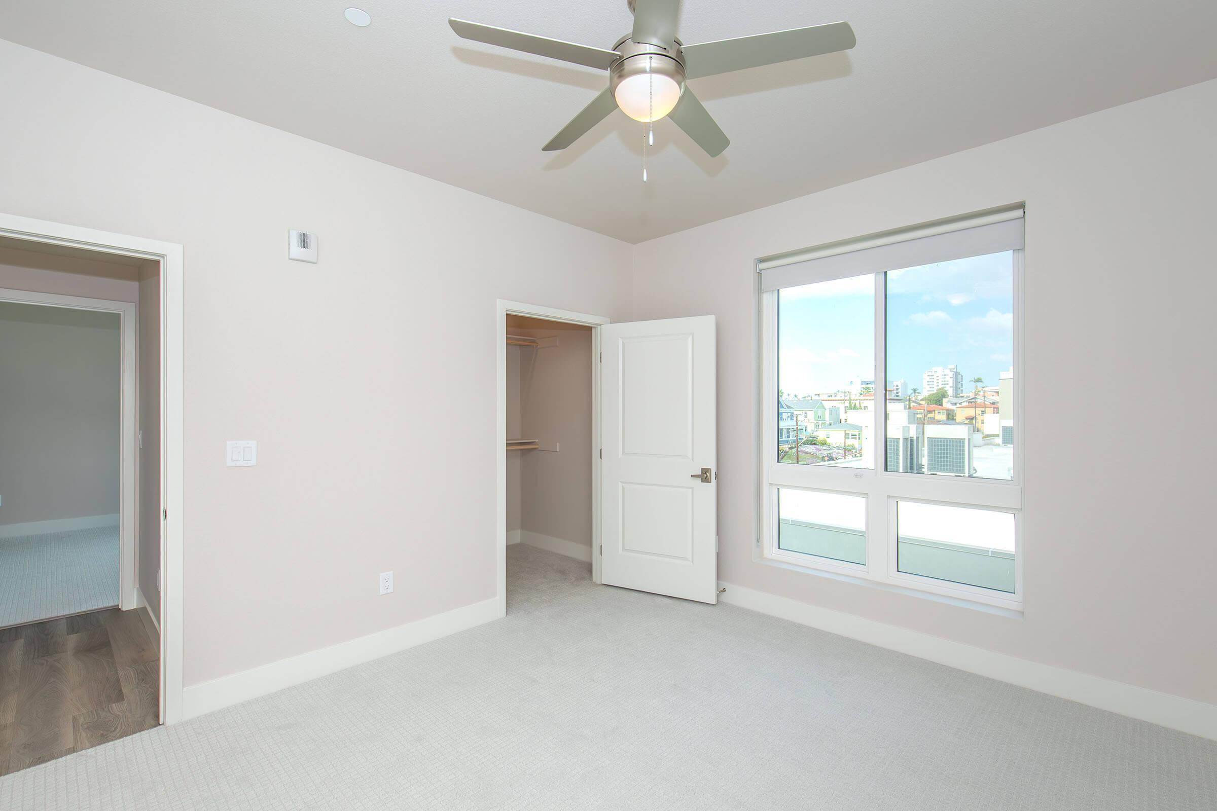 A bright, empty bedroom featuring light-colored walls, a ceiling fan, and a window with city views. The room has light carpet flooring and a door leading to a closet. Another doorway can be seen on the left side, leading to an adjacent room. Natural light fills the space, creating an open and airy feel.