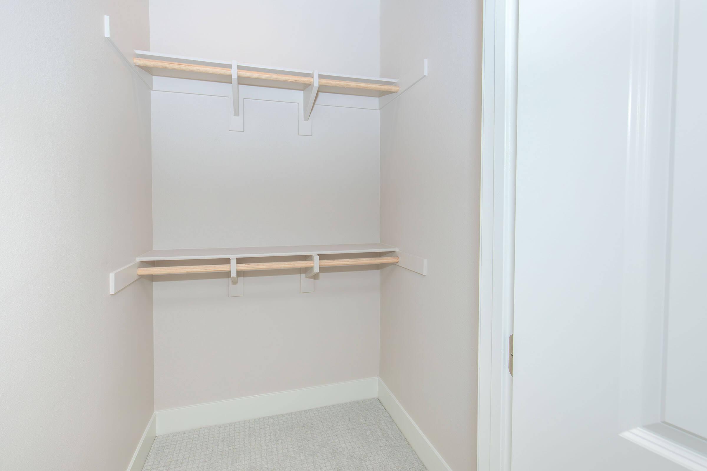 Empty closet with light gray walls and two wooden shelves mounted on white brackets, showcasing ample space for storage. A light gray floor is visible, and a white door partially closed is located on the right side of the image. The overall ambiance is clean and uncluttered.