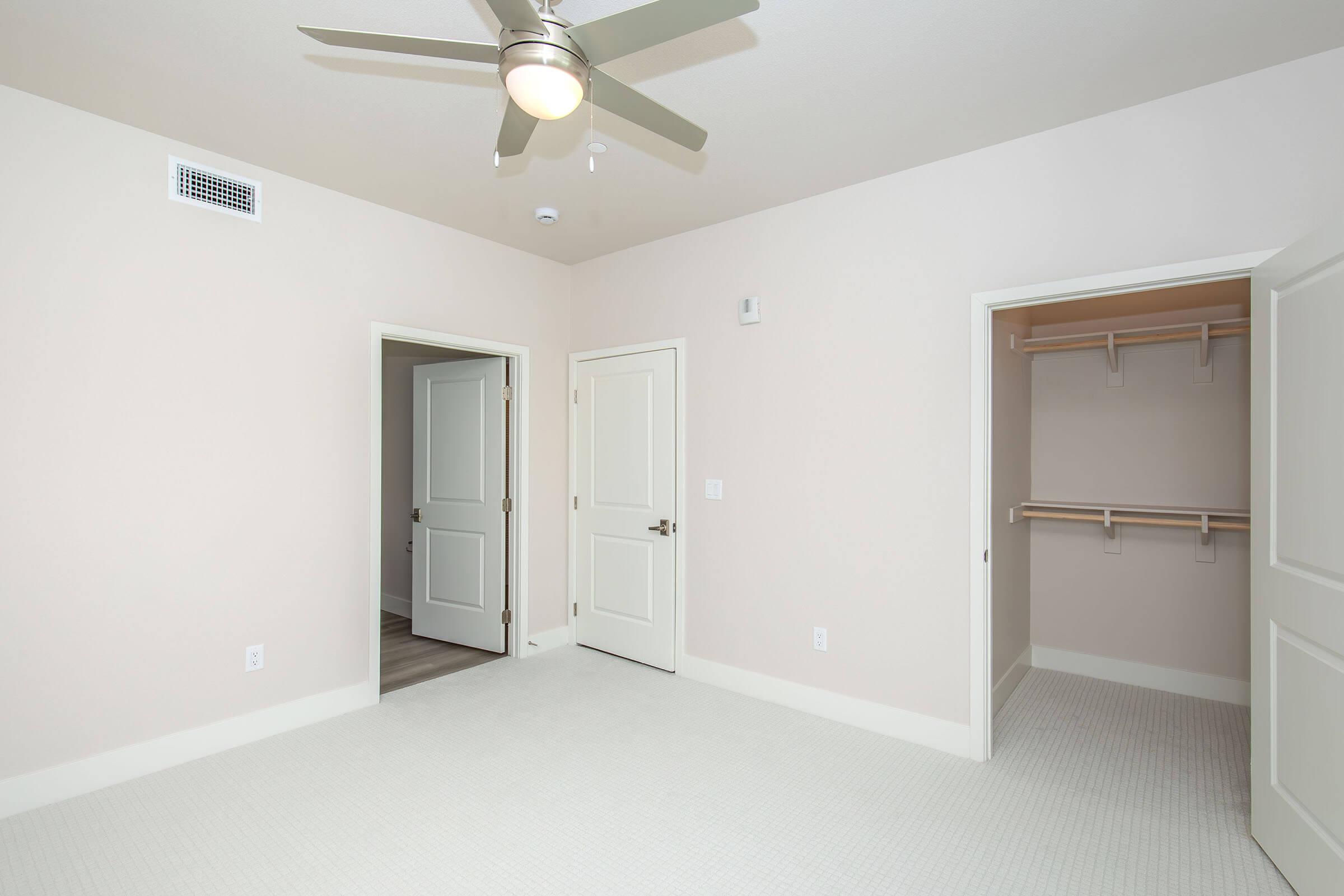 A clean, empty room with light beige walls, a ceiling fan, and carpeted flooring. Two doors are visible: one leading to another room and the other to a spacious closet with shelving. Natural light is present, creating a bright and airy atmosphere.