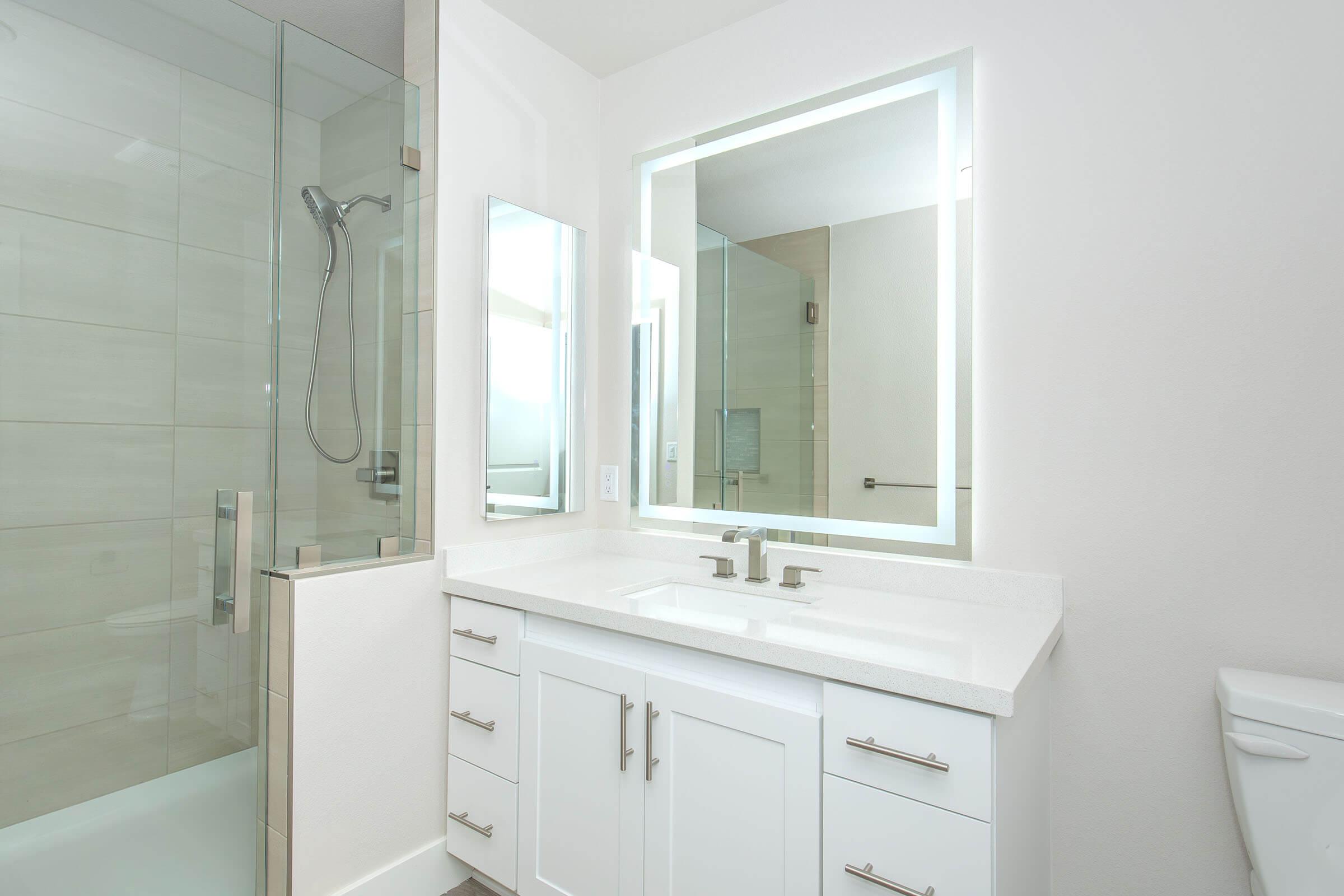 Modern bathroom featuring a sleek white vanity with dual sinks and chrome faucets, illuminated mirror, and a walk-in shower with glass doors. Neutral-colored tile walls create a clean and contemporary look, while a toilet is positioned to the side.