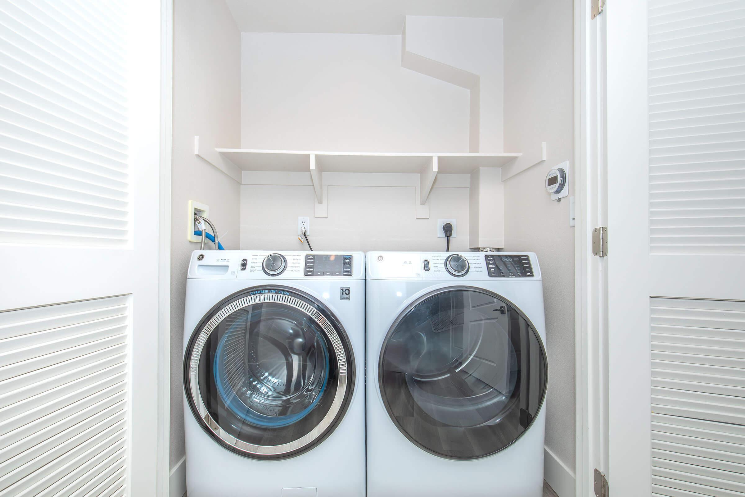 A tidy laundry room featuring a side-by-side washing machine and dryer with clear glass doors, set against a light-colored wall. Above the machines, there is a simple shelf for storage. The space is clean and well-organized, with a closed door in the view.