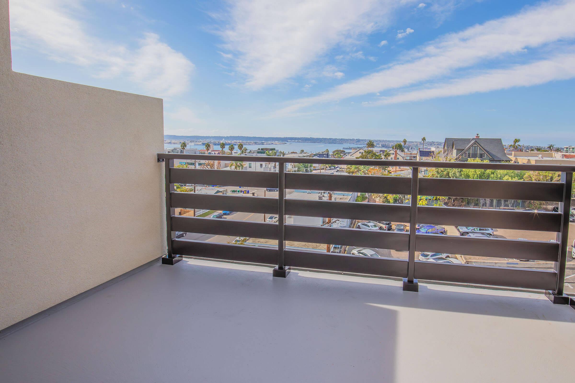 A view from a balcony overlooking a coastal area with palm trees and parked cars below. The sky is blue with some clouds, and water can be seen in the distance, adding a serene atmosphere to the scene. The balcony features a modern railing design.