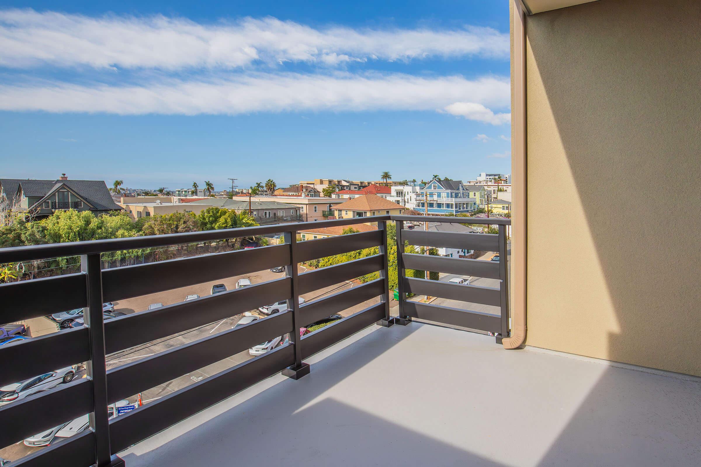 A balcony view overlooking a neighborhood with various houses and a glimpse of the ocean. The scene features blue skies with some clouds, a well-maintained street lined with parked cars, and greenery adding warmth to the urban landscape.
