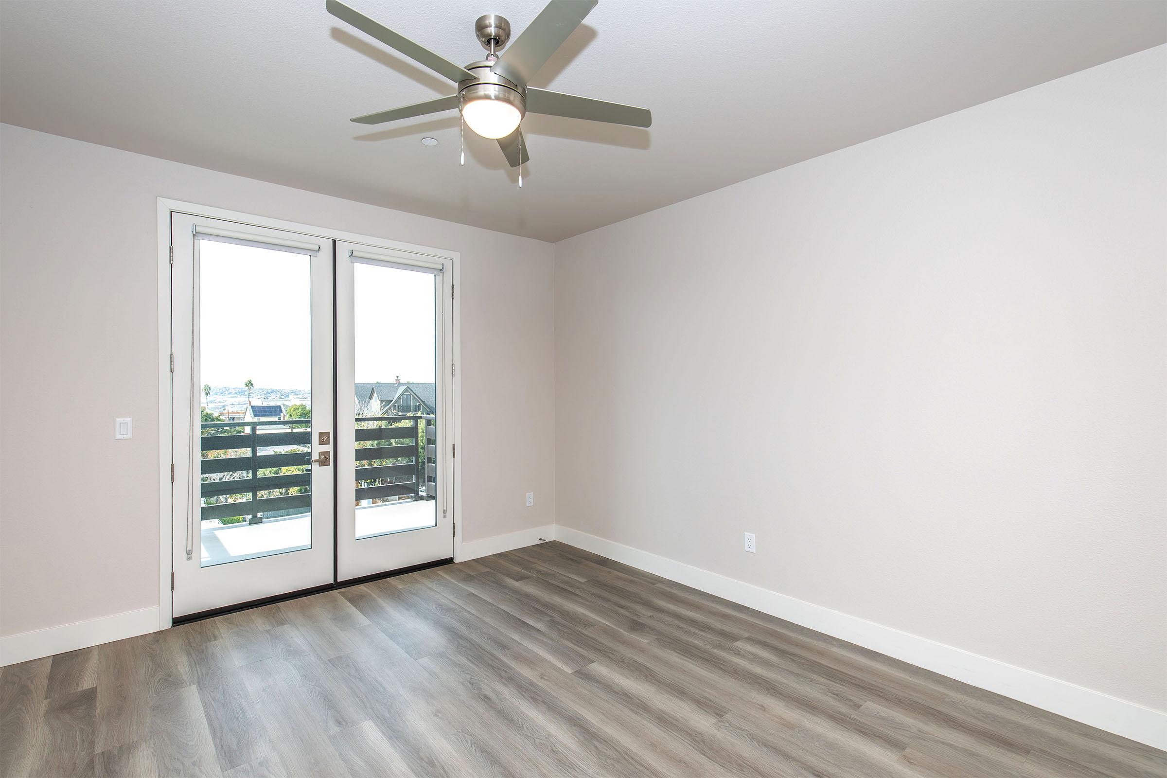 An empty room featuring light gray walls, a ceiling fan, and laminate flooring. There are double doors leading to a balcony, providing a view of the outdoors. The space is bright and airy, suggesting a modern design with ample natural light.