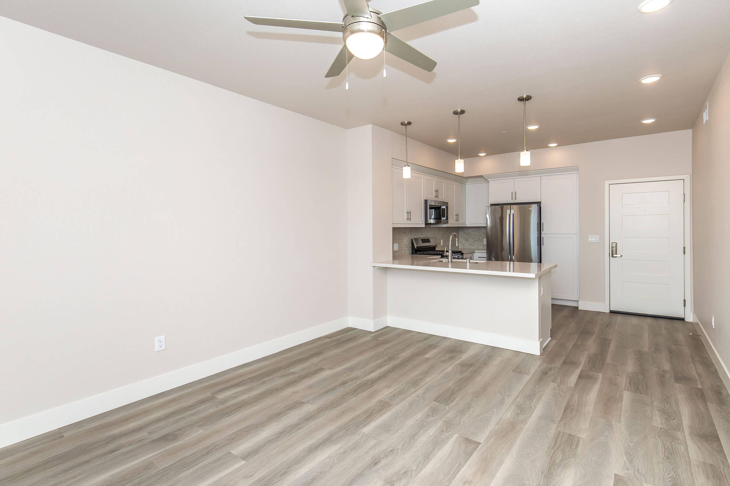 A modern, open-concept living space featuring light-colored walls, wood-style flooring, and a ceiling fan. The kitchen area has stainless steel appliances and pendant lighting. The room offers a spacious layout with a view towards a doorway leading outside.
