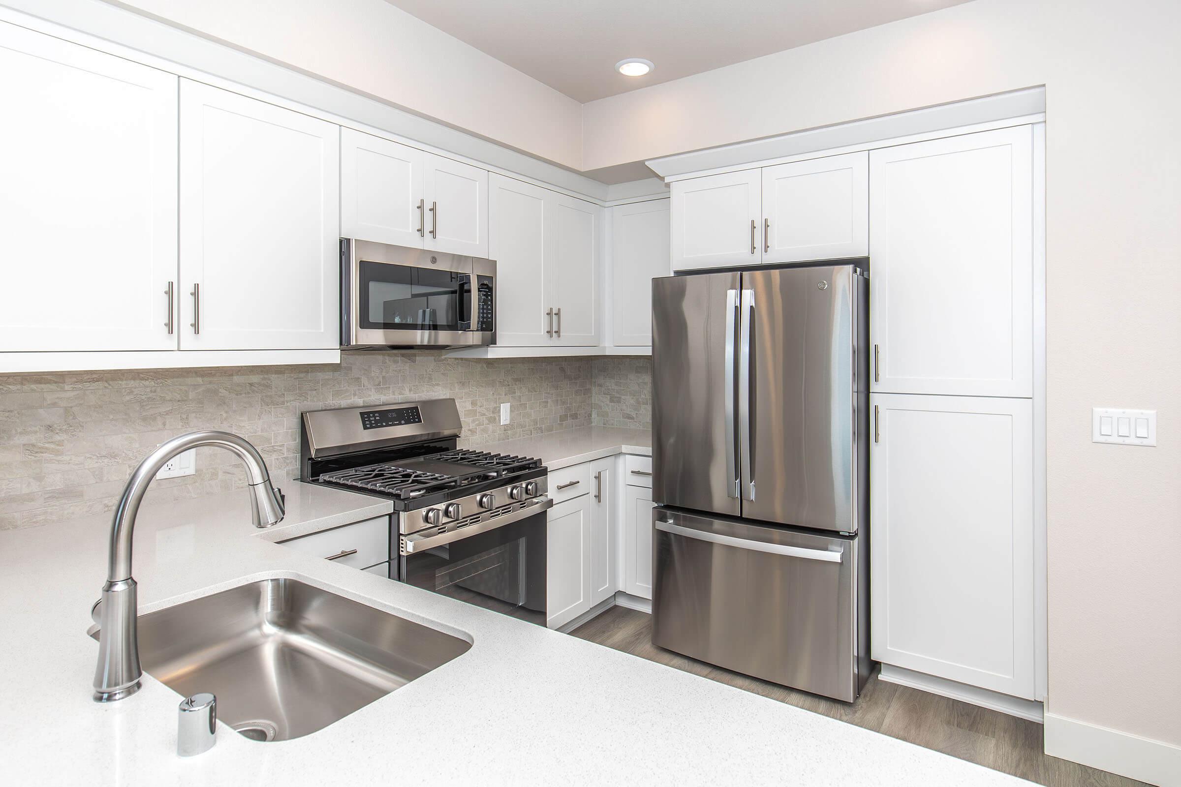 Modern kitchen featuring white cabinetry, stainless steel appliances including a microwave and refrigerator, a gas stove, and a large undermount sink. The countertop is in a light color, and there are neutral-toned tiles on the wall, creating a clean and contemporary aesthetic.