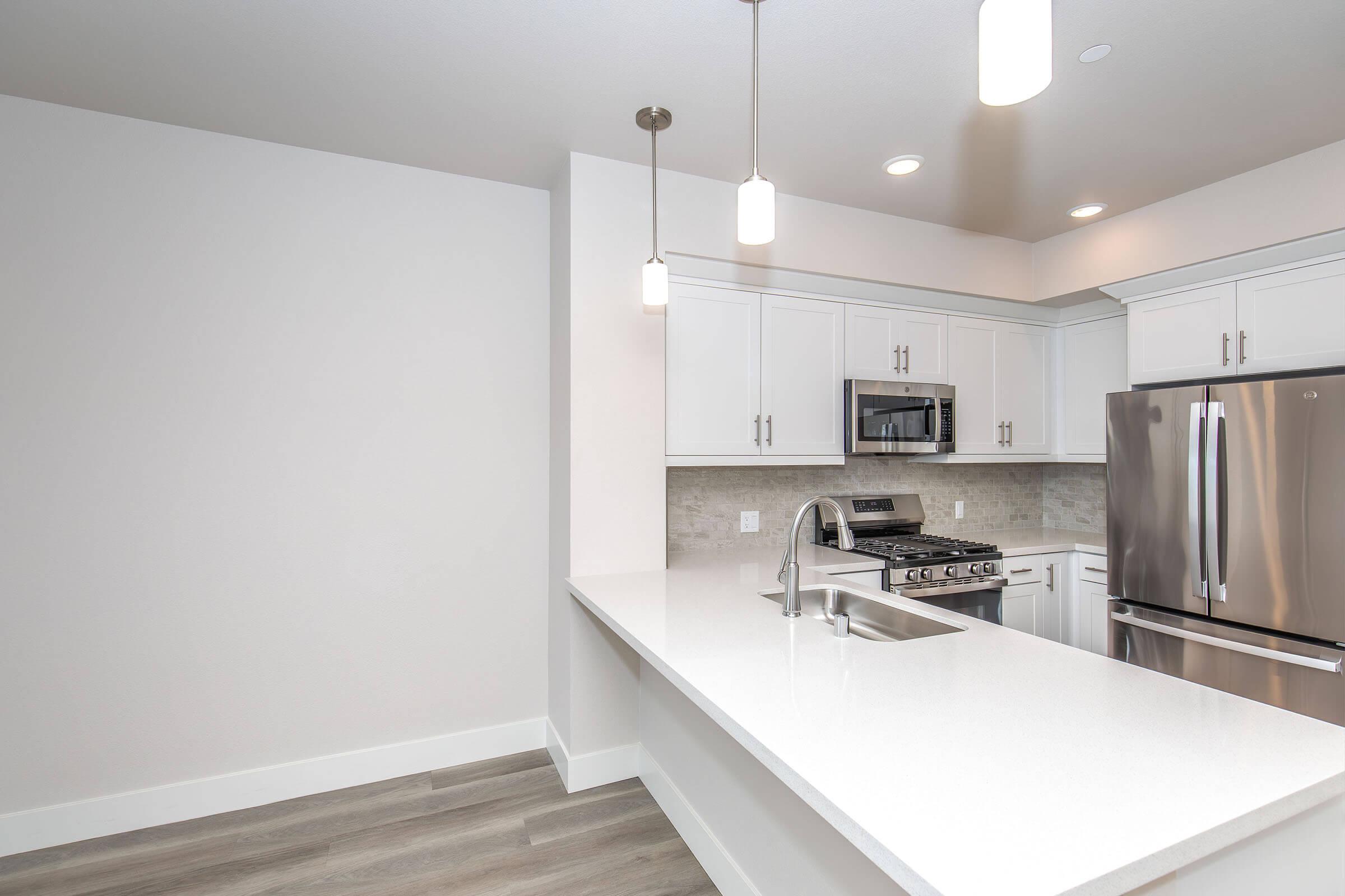 Modern kitchen featuring white cabinetry, stainless steel appliances, and a large island with a sink. Pendant lighting illuminates the space, and the floor is finished with light-colored wood. The design is clean and contemporary, creating an inviting atmosphere.