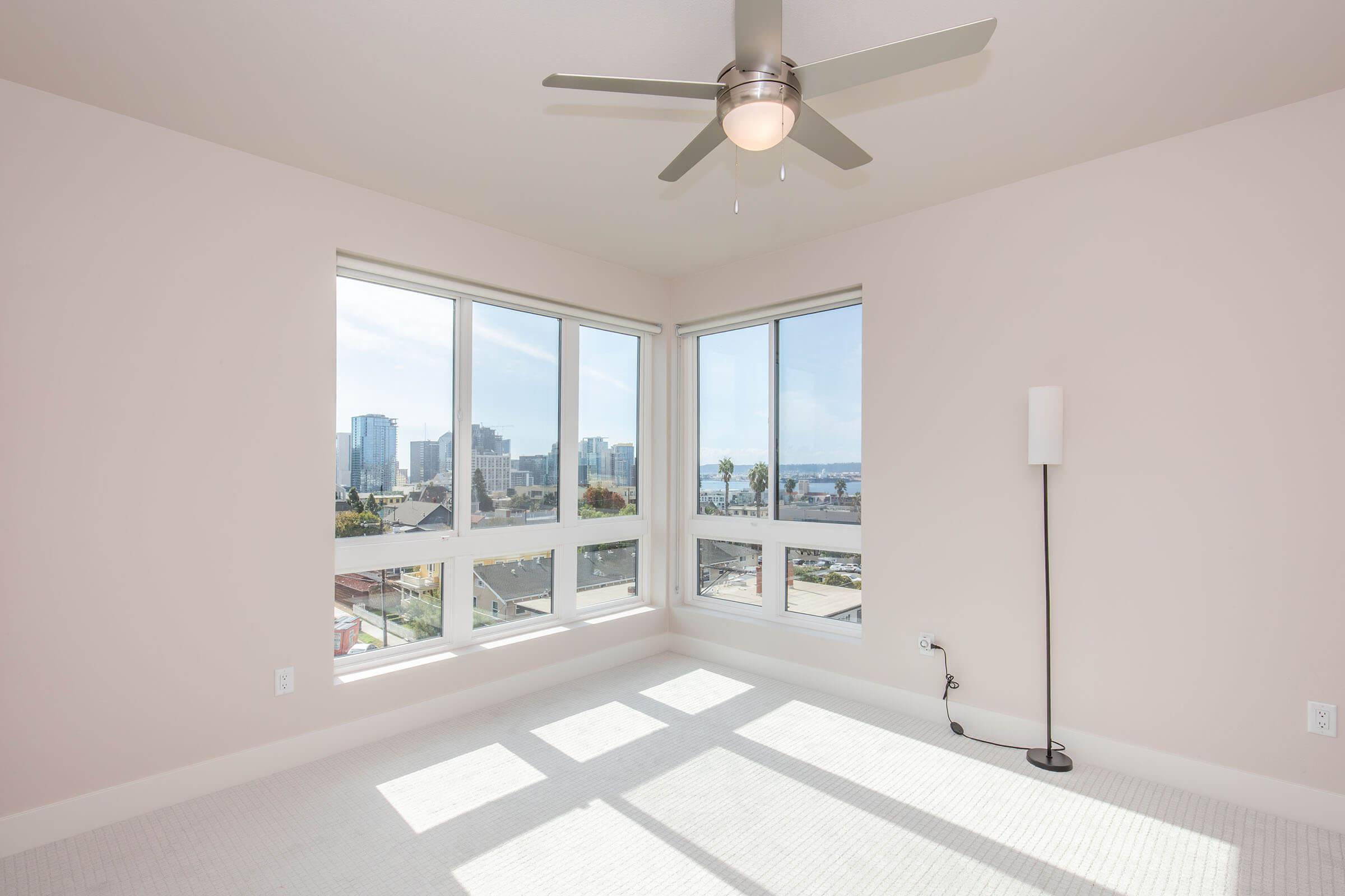 A bright, empty bedroom featuring large windows with a view of a city skyline. The room has light-colored walls, a ceiling fan, and a modern floor lamp. Natural light floods the space, highlighting the clean, minimalistic design.