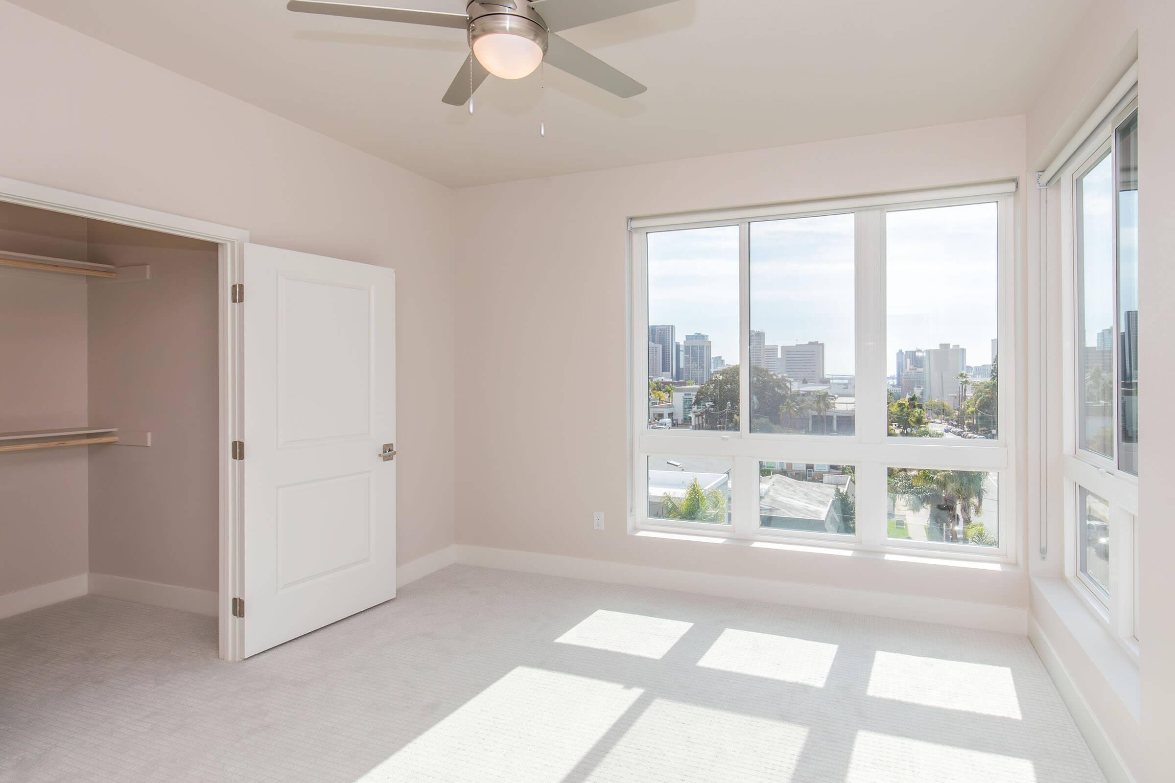 A bright and spacious bedroom featuring a ceiling fan, large windows with a city view, and a closet with an open door. The walls are painted in a light color, and the room has carpet flooring with natural light streaming in.