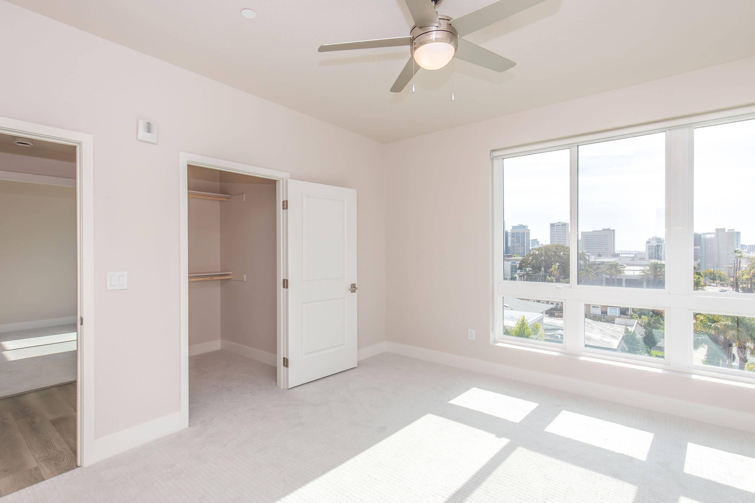 Bright and airy bedroom with light-colored walls, a ceiling fan, and large windows offering a view of the city skyline. The room features a spacious closet with shelving and soft carpet flooring, creating a welcoming atmosphere. Natural light streams in through the windows.