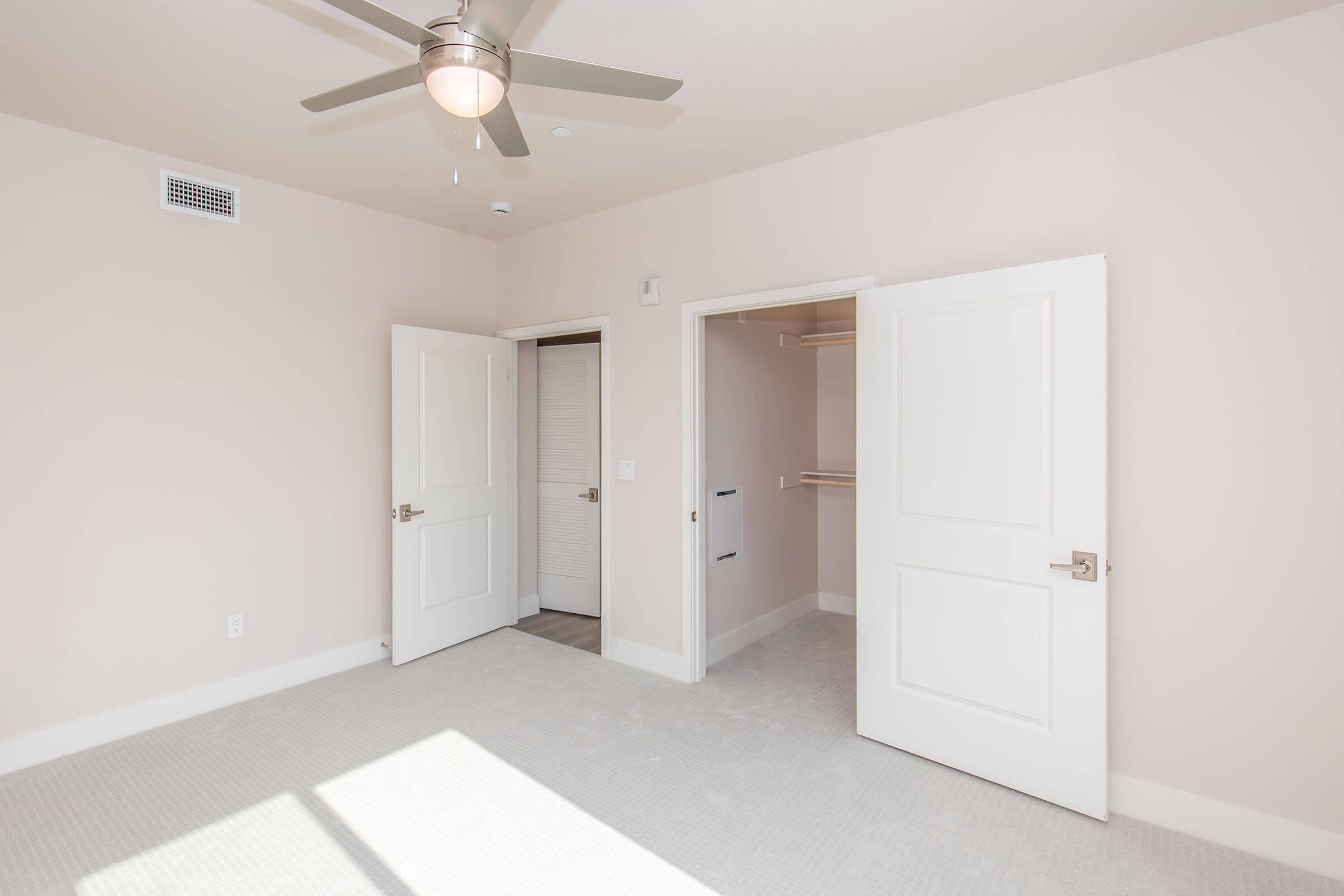 A well-lit, empty room featuring light beige walls, a ceiling fan, and two white doors. One door leads to a closet with shelving. The floor is carpeted in a light shade, creating a spacious and airy atmosphere.