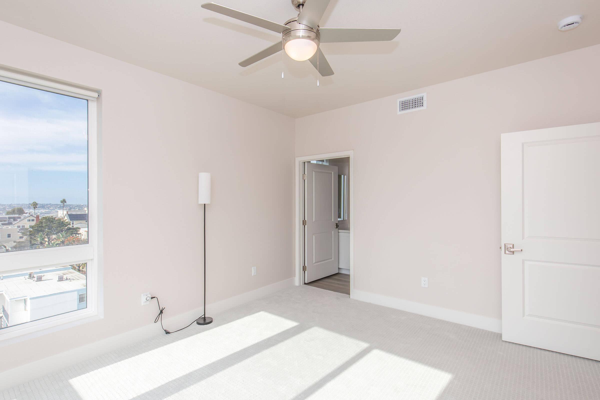 An empty room with a ceiling fan, large window allowing natural light, and light-colored walls. A tall lamp stands next to the wall, and an open door leads to another room. The floor is carpeted in a soft, neutral color. The overall atmosphere is bright and airy.