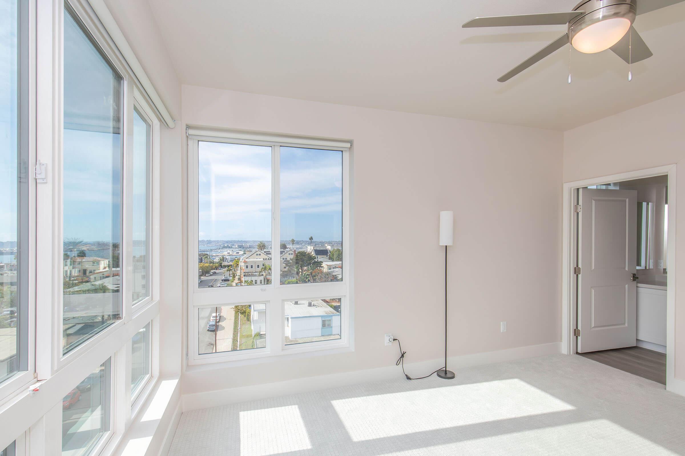 Bright and airy room with large windows offering a view of a sunny exterior, including a nearby harbor and greenery. The space features a light-colored carpet, a modern ceiling fan, and a minimalist design with a lamp and an open doorway leading to another area, creating a welcoming atmosphere.