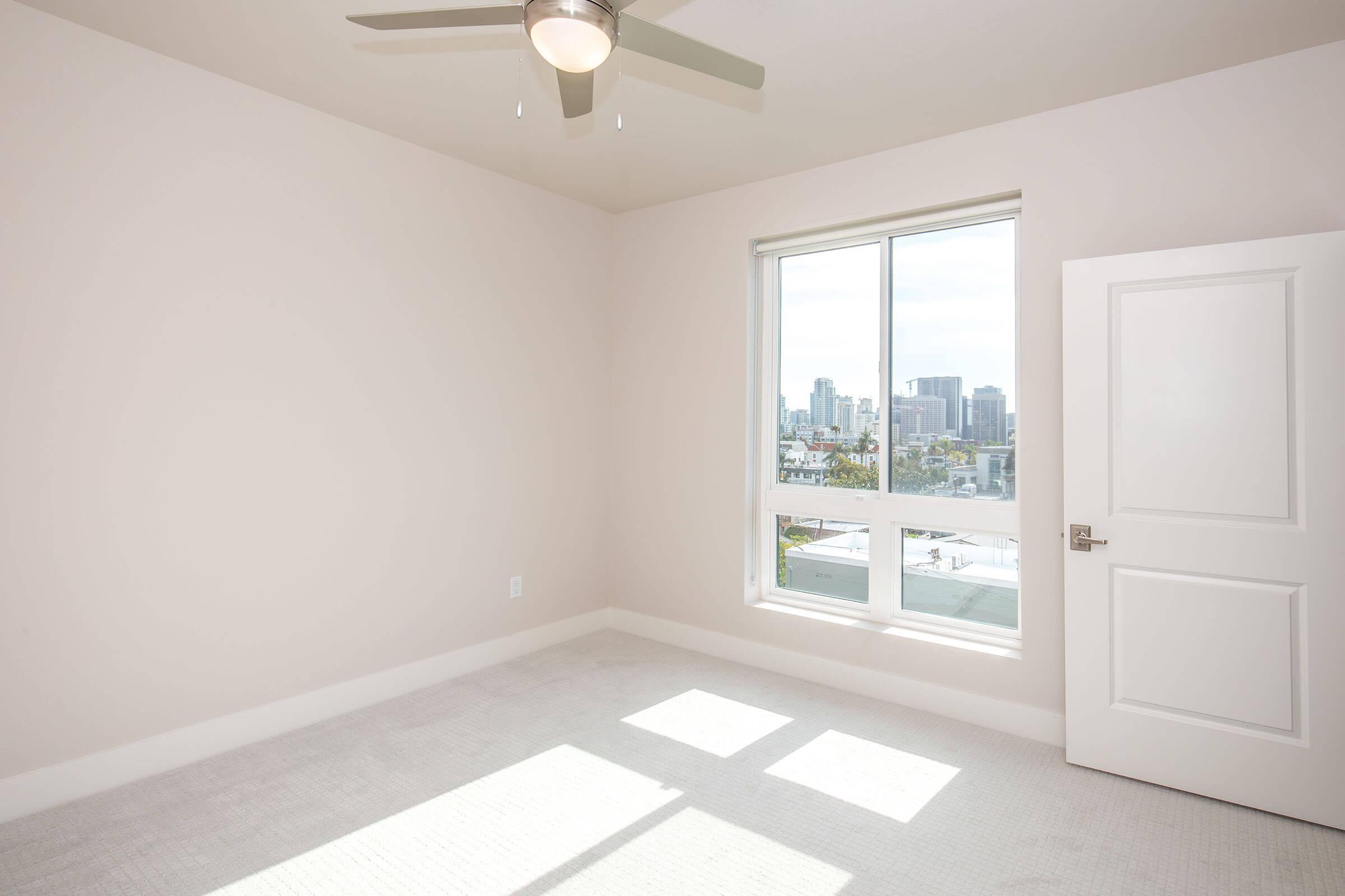 A bright, empty bedroom featuring light-colored walls and carpet. A window allows natural light to fill the space, showcasing a city skyline in the background. A ceiling fan is visible, and a white door leads to another area. The room is clean and uncluttered, ready for new occupants.