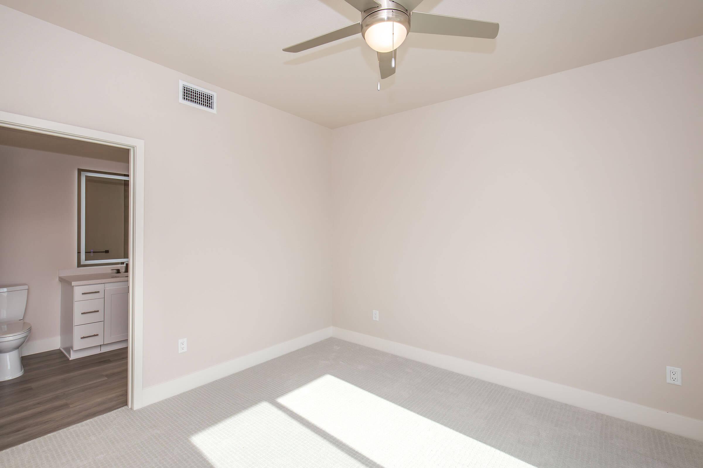 A spacious, empty bedroom featuring light-colored walls and carpet. A ceiling fan is installed, and there is a doorway leading to an adjoining bathroom with a toilet and vanity. Natural light enters through a window, creating a bright atmosphere in the room.