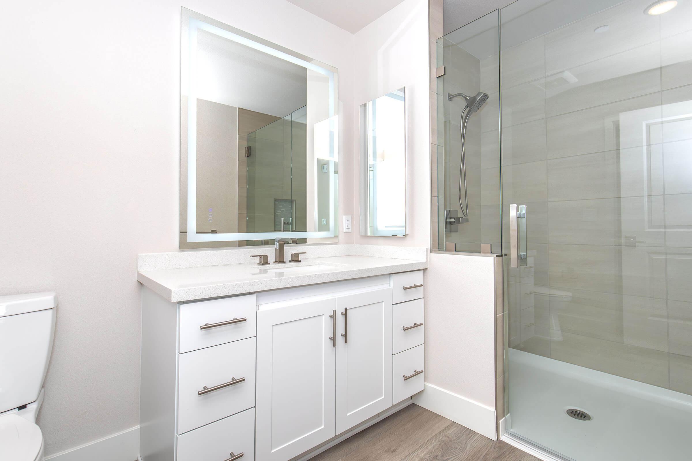 A modern bathroom featuring a double sink vanity with white cabinetry, silver handles, and a large mirror. To the right, there is a walk-in shower with glass doors and neutral tiled walls. The flooring is light wood, creating a bright and spacious atmosphere.