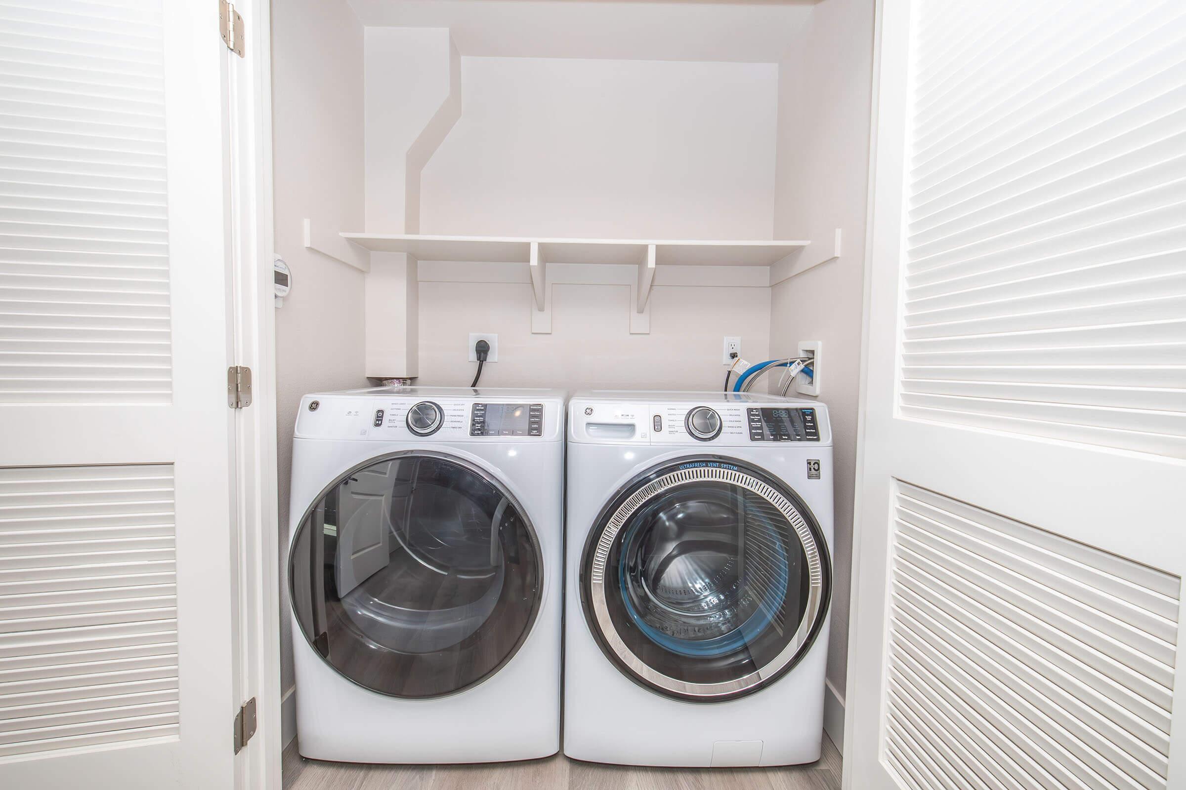 A laundry room with two modern, front-loading washing machines side by side. The machines are white and have large glass doors. Above them is a shelf for storage. The room has light-colored walls and a sleek, clean aesthetic.