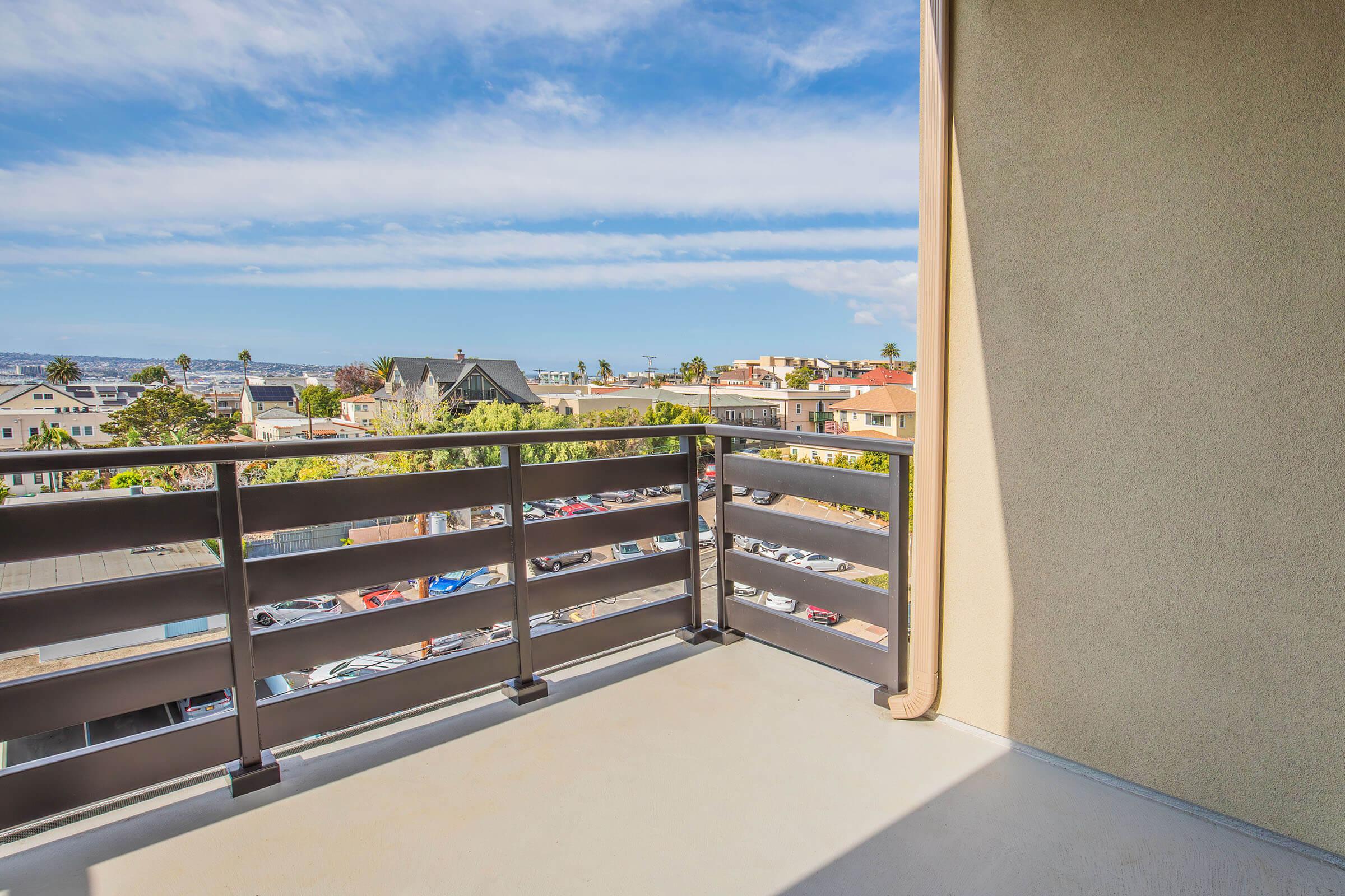 A balcony view overlooking a coastal neighborhood with houses and parked cars. The sky is partly cloudy, creating a bright and inviting atmosphere. The railing of the balcony frames the scene, highlighting the peaceful surroundings.
