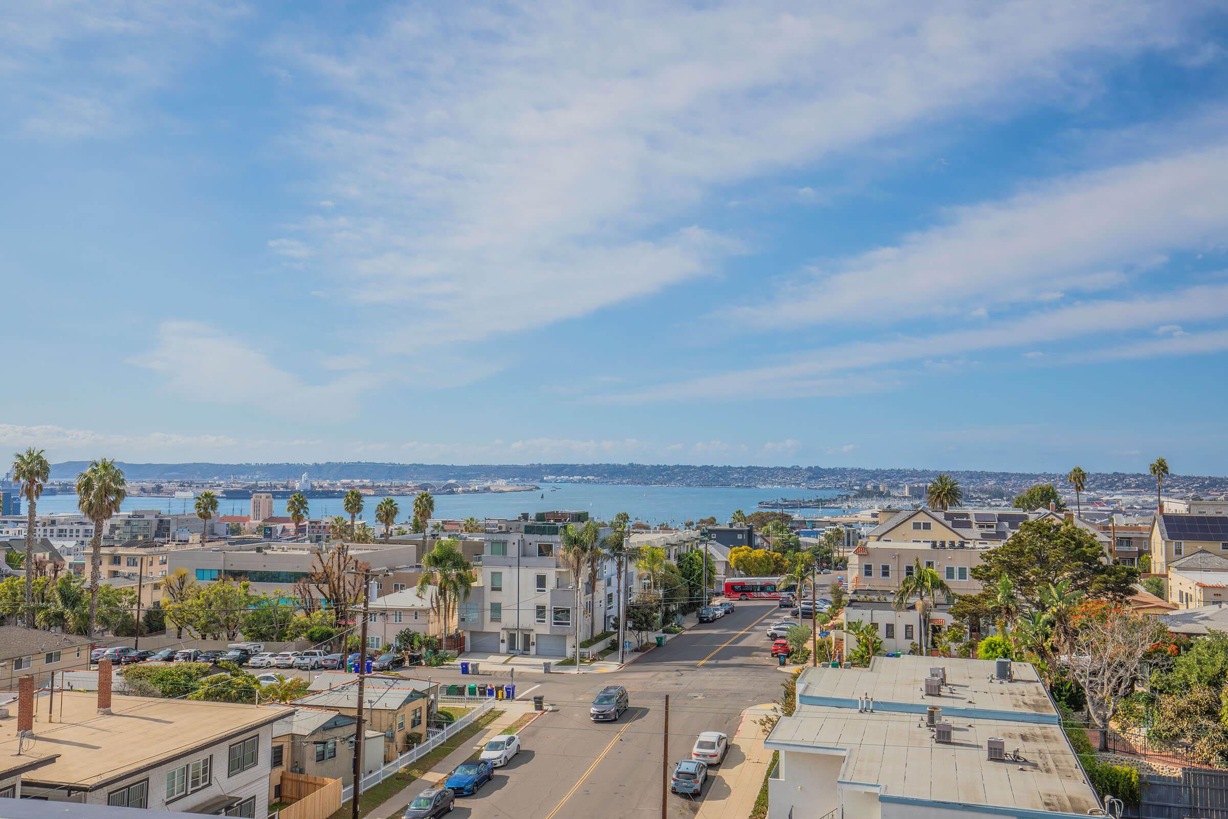 A scenic view of a coastal city with a mix of residential buildings and palm trees. The skyline is partially obscured by clouds under a bright blue sky, and distant hills are visible across the bay. Streets with parked cars run through the foreground, creating a vibrant urban atmosphere near the water.