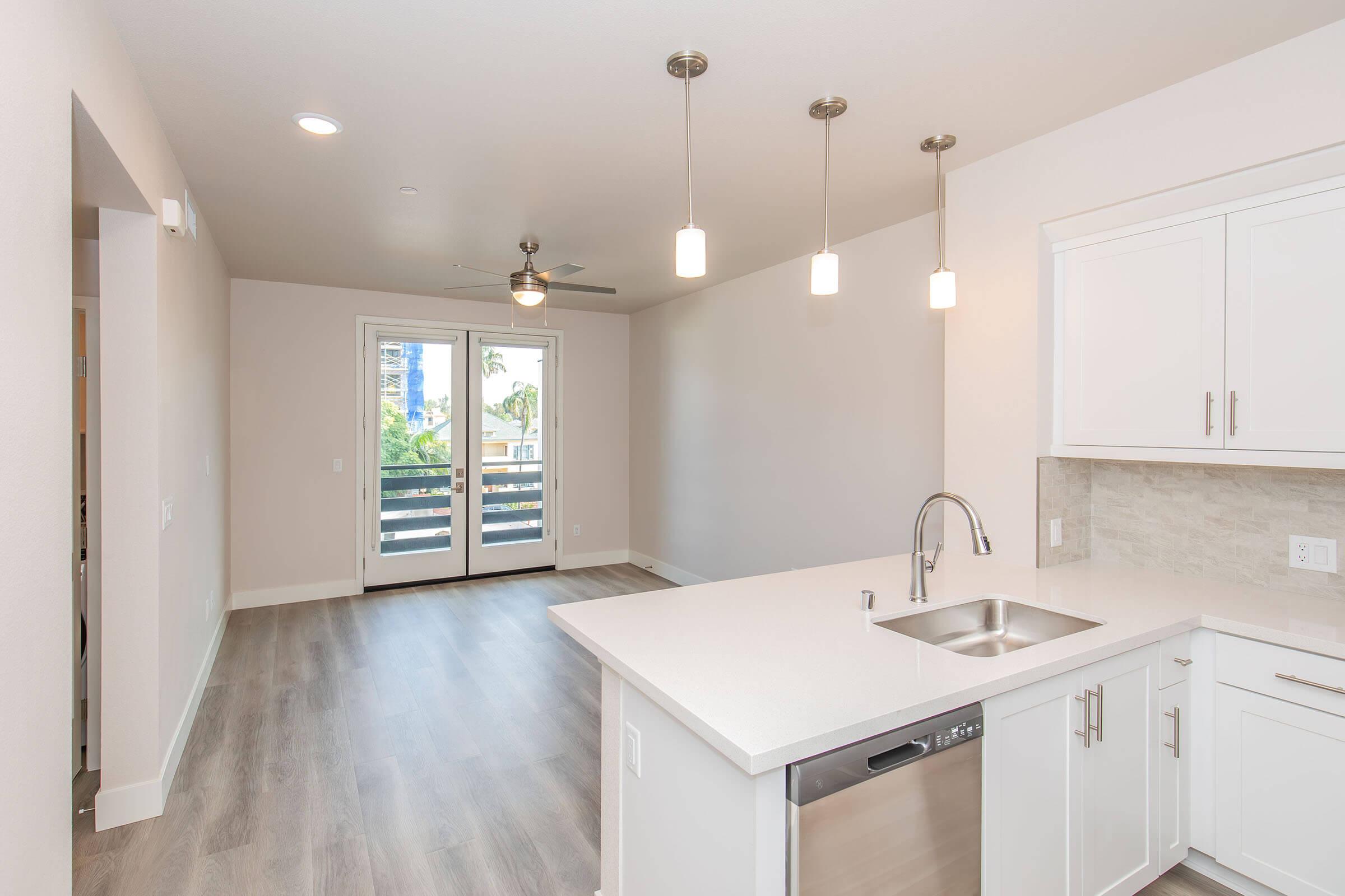 A modern kitchen with a white countertop and stainless steel sink, featuring a dishwasher. The kitchen opens to a spacious living area with light-colored walls and wood-like flooring. Two glass doors lead to a balcony, offering a view of the outdoors. Three pendant lights hang from the ceiling, providing illumination.