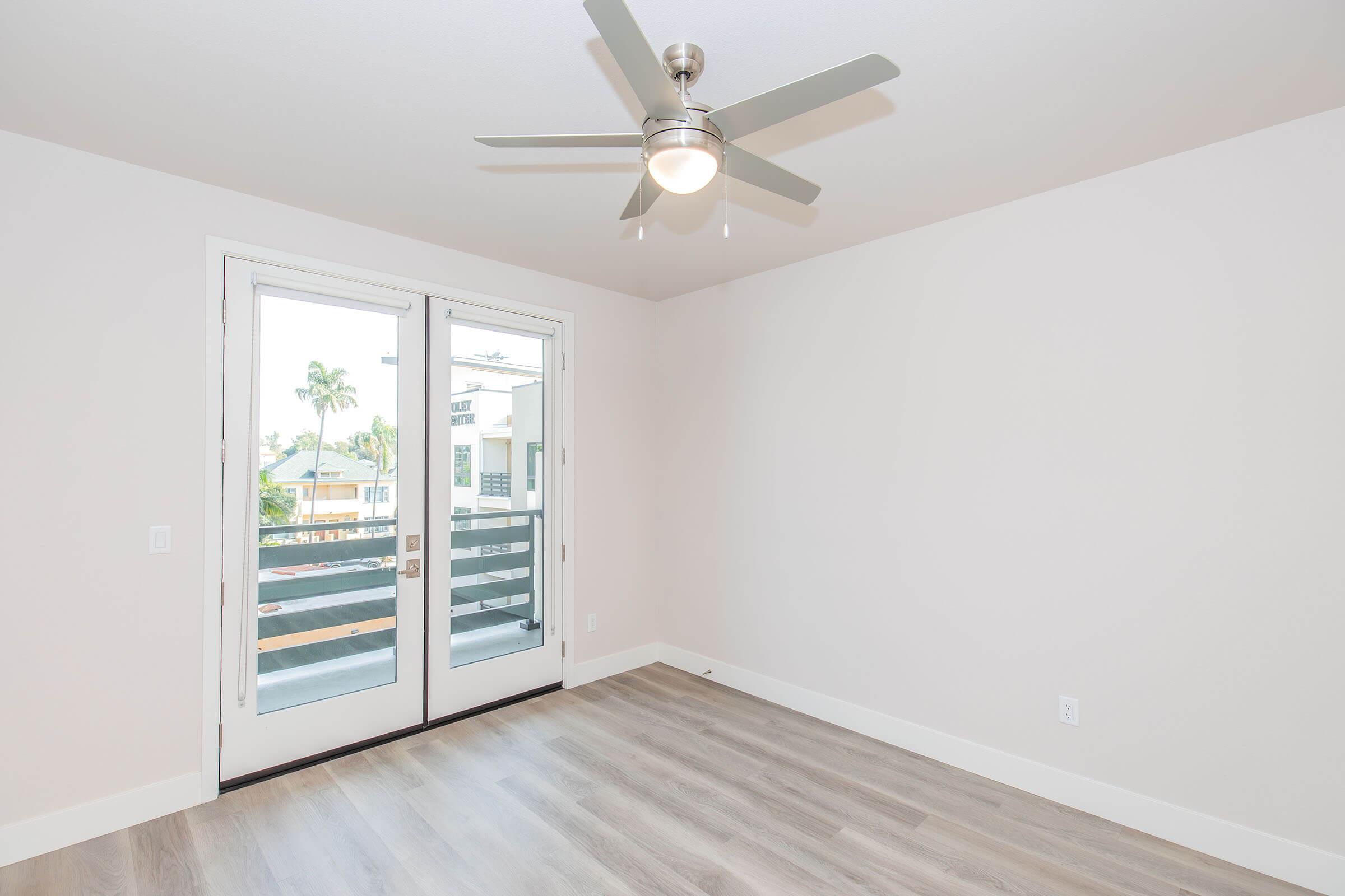 A bright, empty room with light-colored walls and hardwood flooring. It features a modern ceiling fan and large sliding glass doors leading to a balcony. Natural light spills in from outside, showcasing a glimpse of palm trees in the distance.