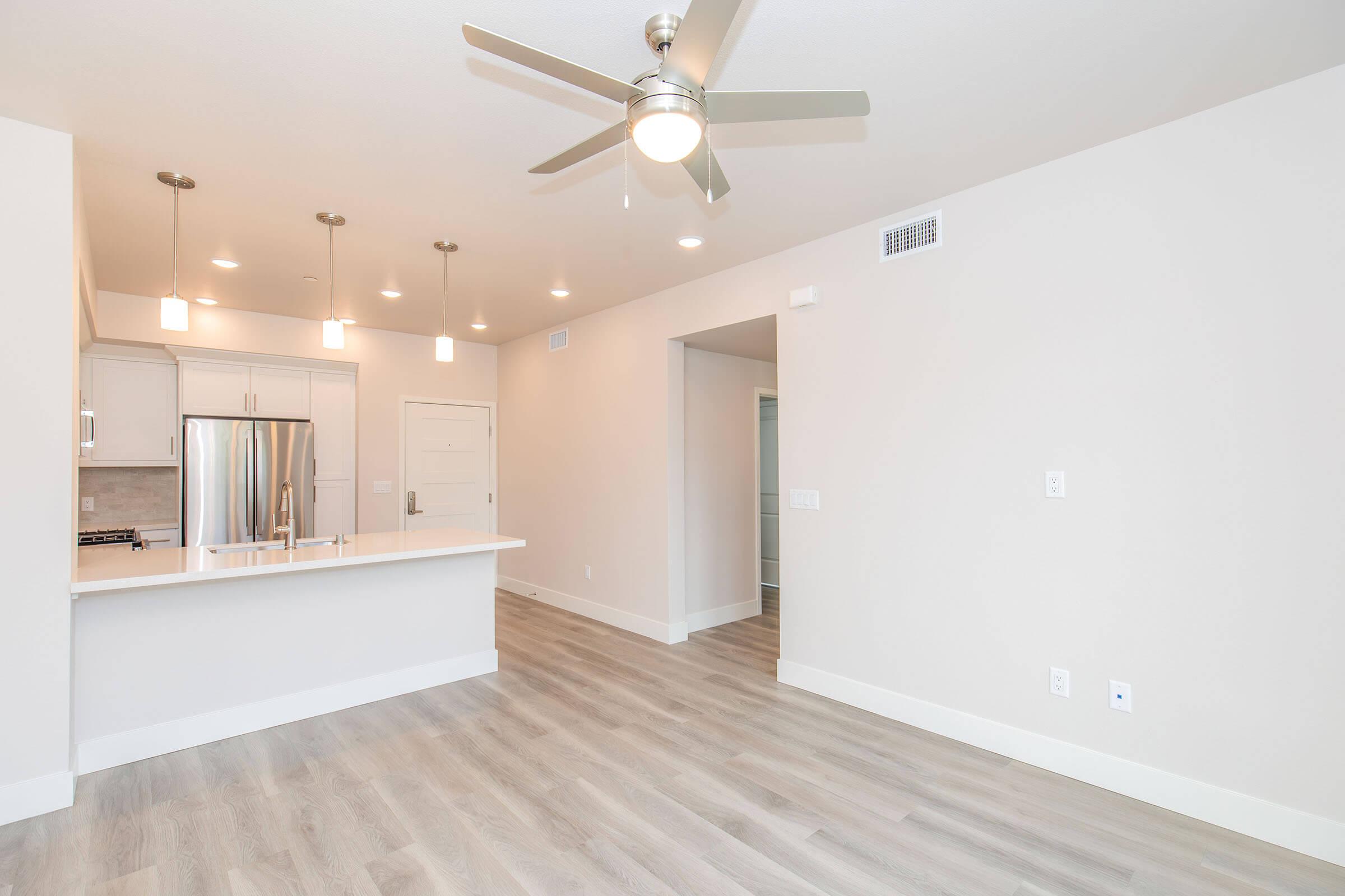 Modern open-concept kitchen and living area with light wood flooring, white walls, and a ceiling fan. The kitchen features stainless steel appliances and a large island with bar seating. Brightly lit with pendant lights, the space is airy and inviting, leading to a doorway at the back.