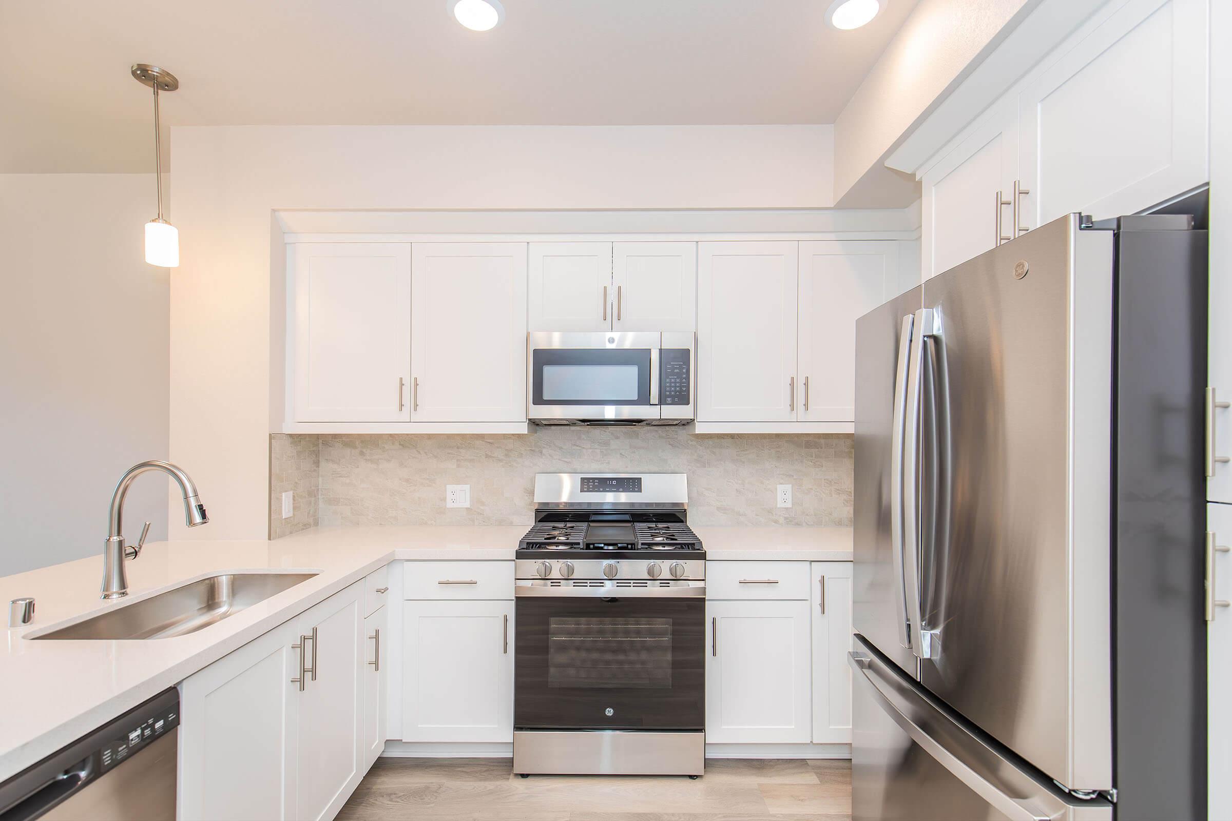 A modern kitchen featuring white cabinetry, a stainless steel gas stove, an over-the-range microwave, and a refrigerator. The countertops are light-colored, with a sink and a single pendant light above. The decor is sleek and contemporary, showcasing a neutral color palette.
