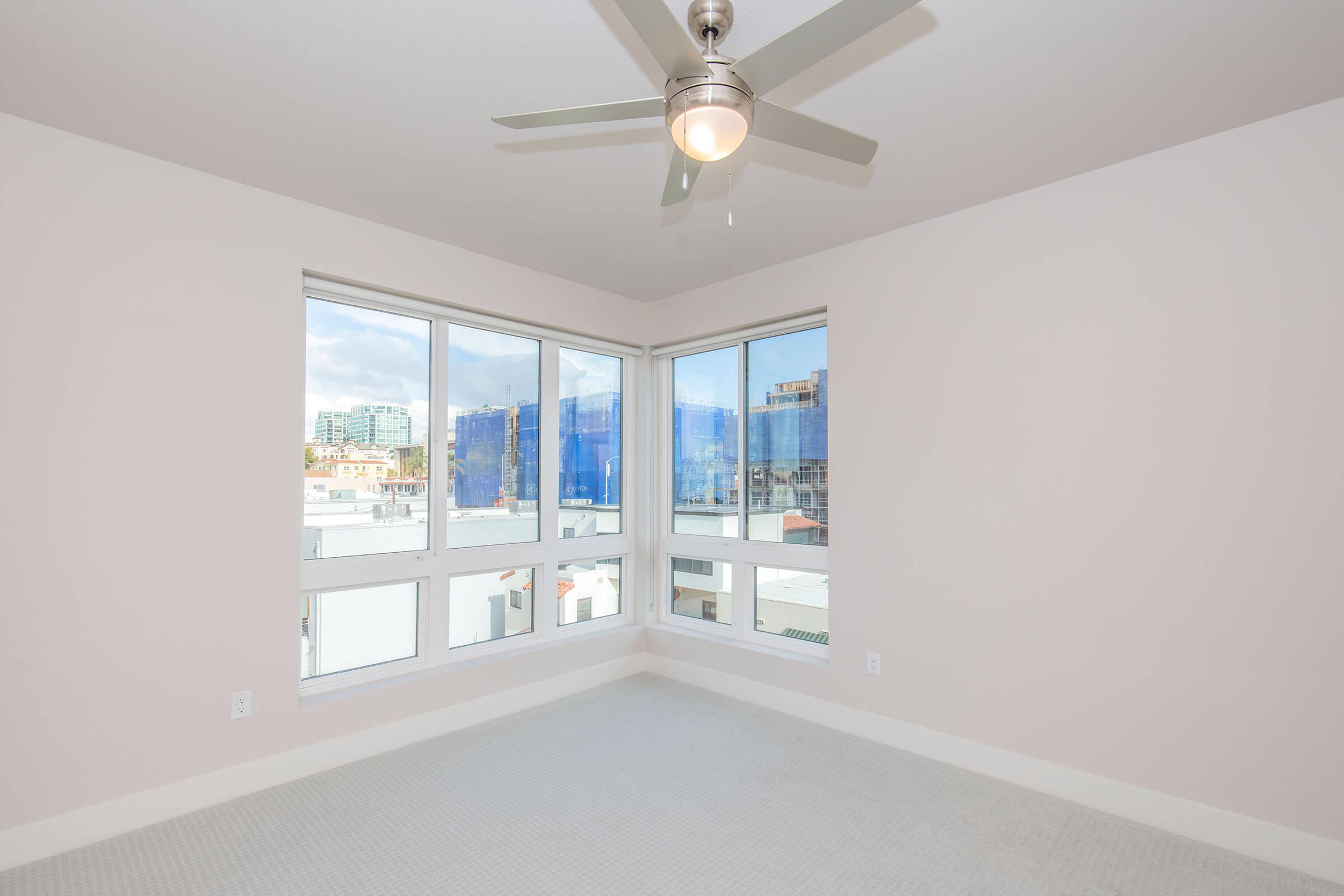 A bright, empty bedroom with light-colored walls and a ceiling fan. Large windows offer a view of a cityscape with blue construction scaffolding visible outside. The floor is carpeted and the overall atmosphere is open and airy.