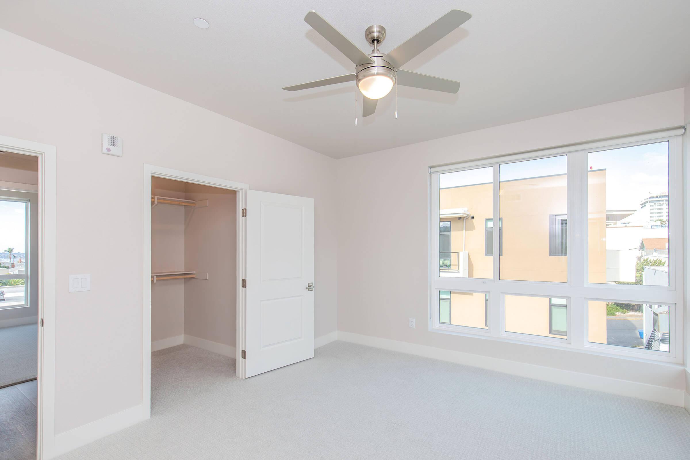 A spacious, light-filled bedroom featuring a ceiling fan, large window with a view, and a walk-in closet. The walls are painted a soft neutral color, and there's plush carpet on the floor, creating a comfortable and inviting atmosphere.