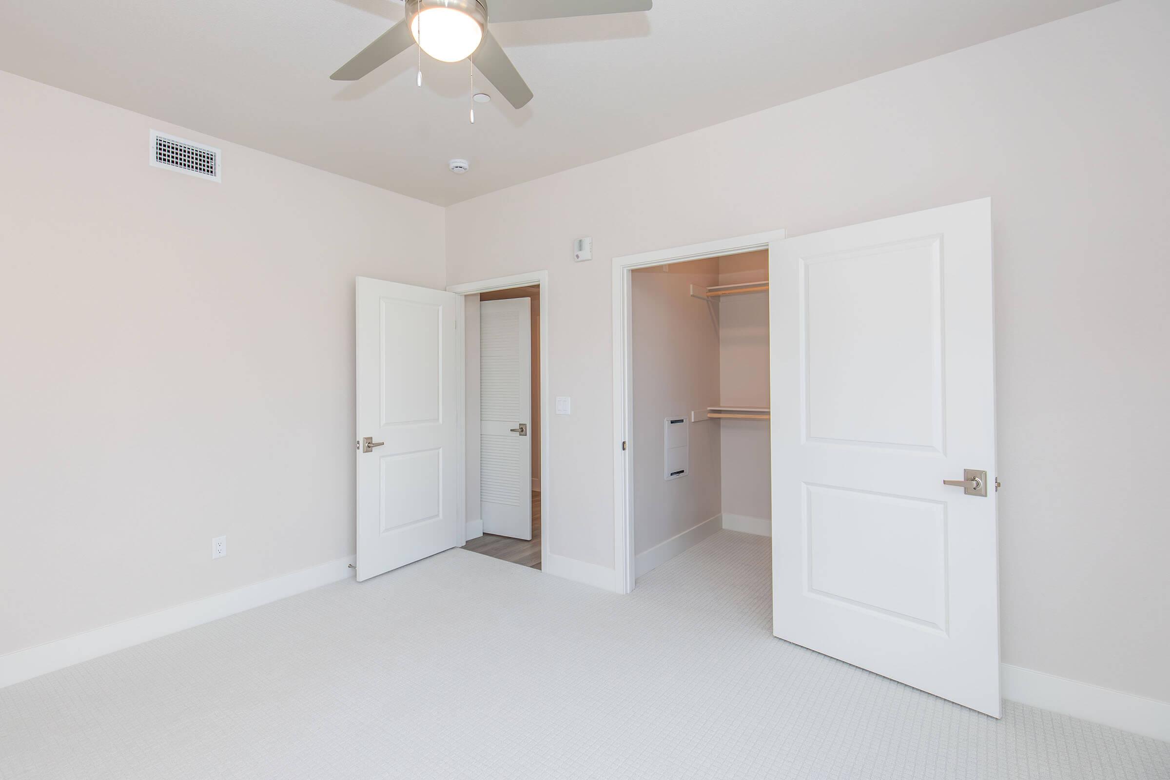 A bright, empty bedroom featuring light-colored walls and carpet. Two open doors lead to a closet and a hallway. A ceiling fan is visible above, enhancing the spacious feel of the room. The overall atmosphere is clean and modern, suitable for personalization.