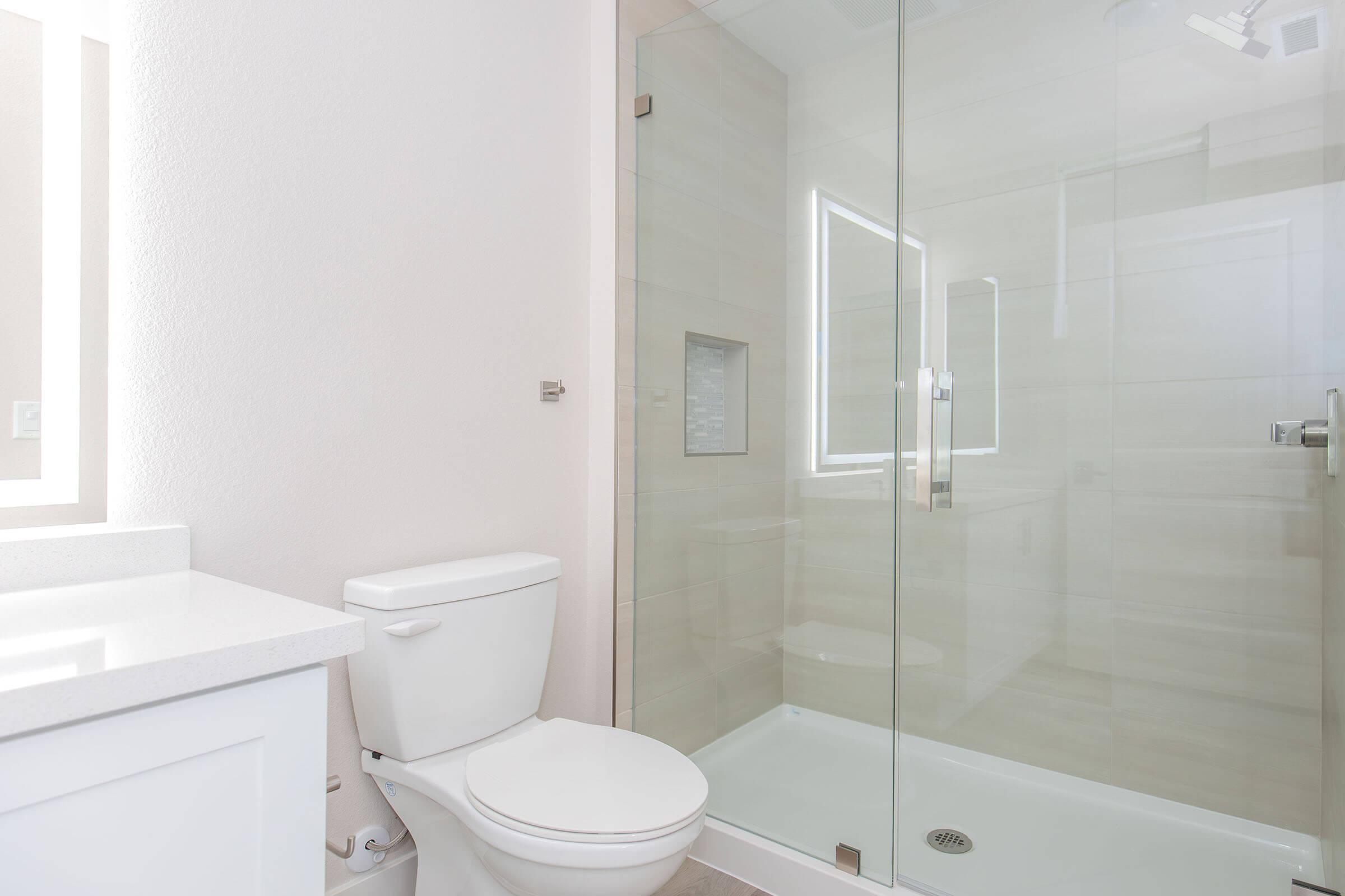 Modern bathroom interior featuring a glass shower stall, a white toilet, and a sleek vanity with a countertop. Soft beige tiles adorn the walls, creating a clean and bright atmosphere, complemented by natural light. The design emphasizes simplicity and functionality.
