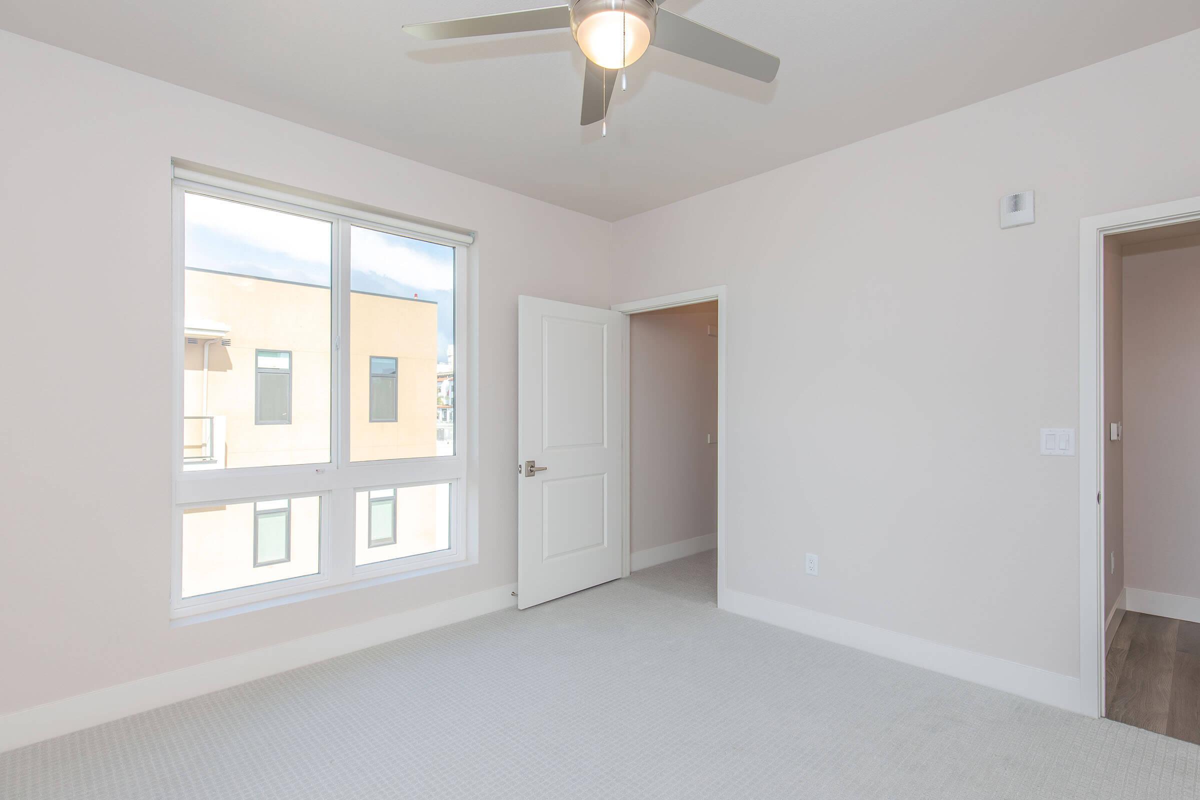 A bright, empty room featuring light-colored walls and carpet, with a large window allowing natural light and a ceiling fan. The open door leads to another space, and there's a small wall vent visible. The overall ambiance is clean and modern, ideal for various decorative possibilities.
