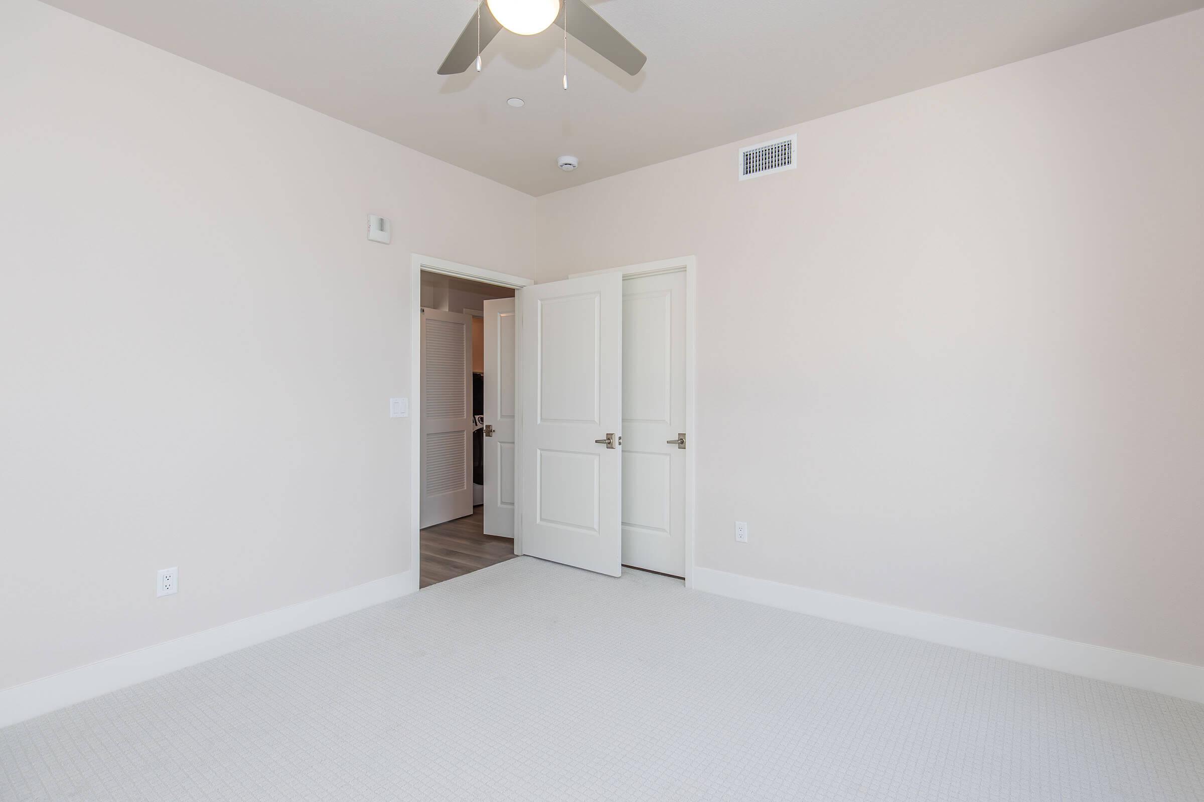 A spacious, empty room with light beige walls and carpet. A ceiling fan is mounted above, and there is an open double door leading to another room. Natural light illuminates the space, creating a bright and inviting atmosphere.