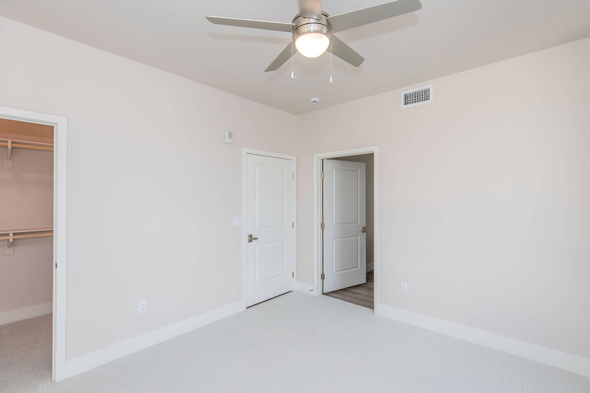Interior of a light-colored room featuring a ceiling fan, two doors leading to separate areas, and a spacious closet. The flooring is carpeted, and the walls are painted in neutral tones, creating a bright and airy atmosphere.