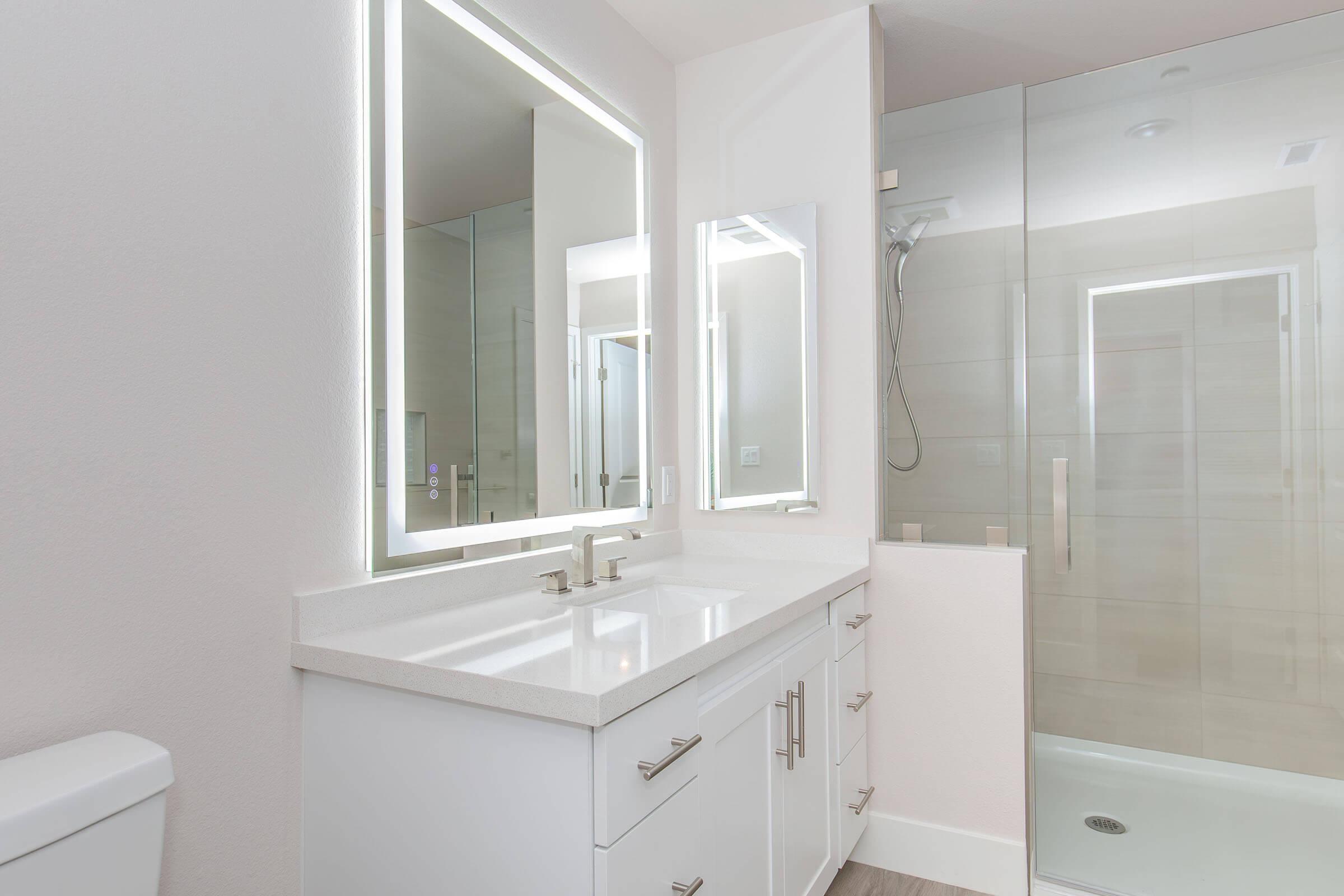 Modern bathroom featuring a dual sink vanity with a white countertop and elegant faucets. A large illuminated mirror above the sinks enhances the bright atmosphere. A glass-enclosed shower with a minimalist design is visible in the background, alongside neutral-colored walls and flooring.