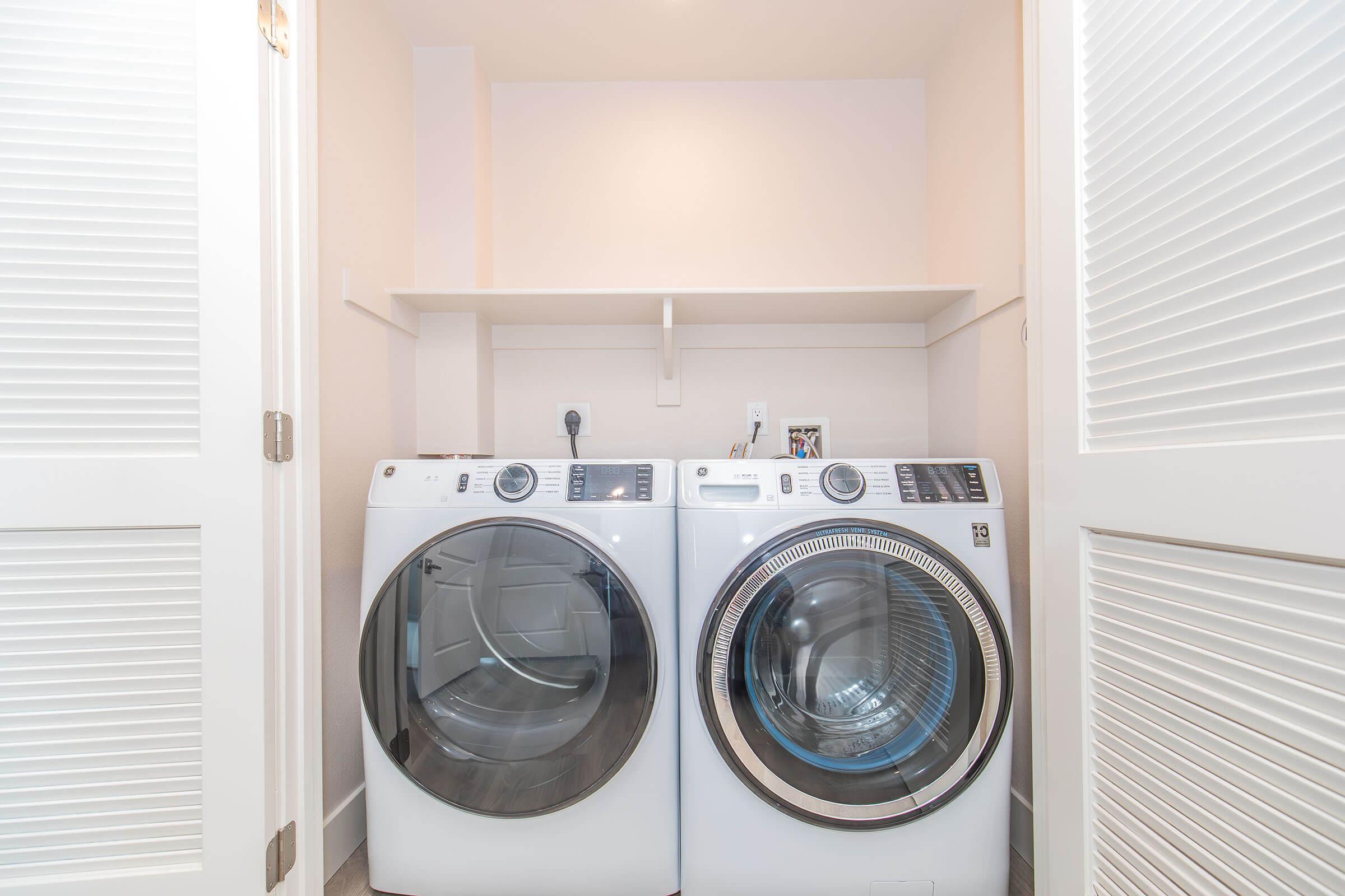 A modern laundry room featuring a stacked washer and dryer in a compact space. The appliances are white with glass doors, surrounded by light-colored walls and shelving above for storage. The room has a clean and organized appearance, emphasizing efficiency in a small area.