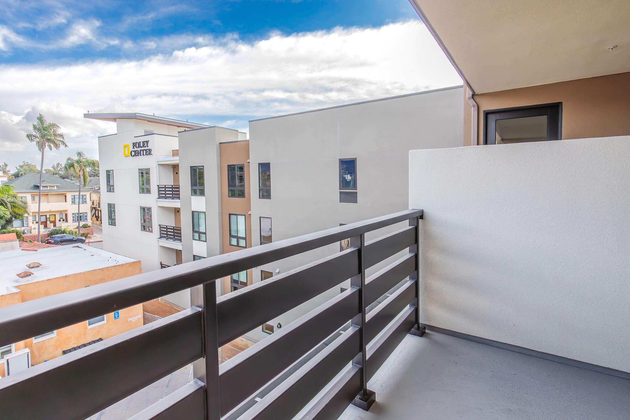 A view from a balcony overlooking a modern urban environment. In the foreground, there is a metal railing, and in the background, there are buildings with flat roofs and large windows. A clear blue sky with some clouds can be seen above, indicating a pleasant day.