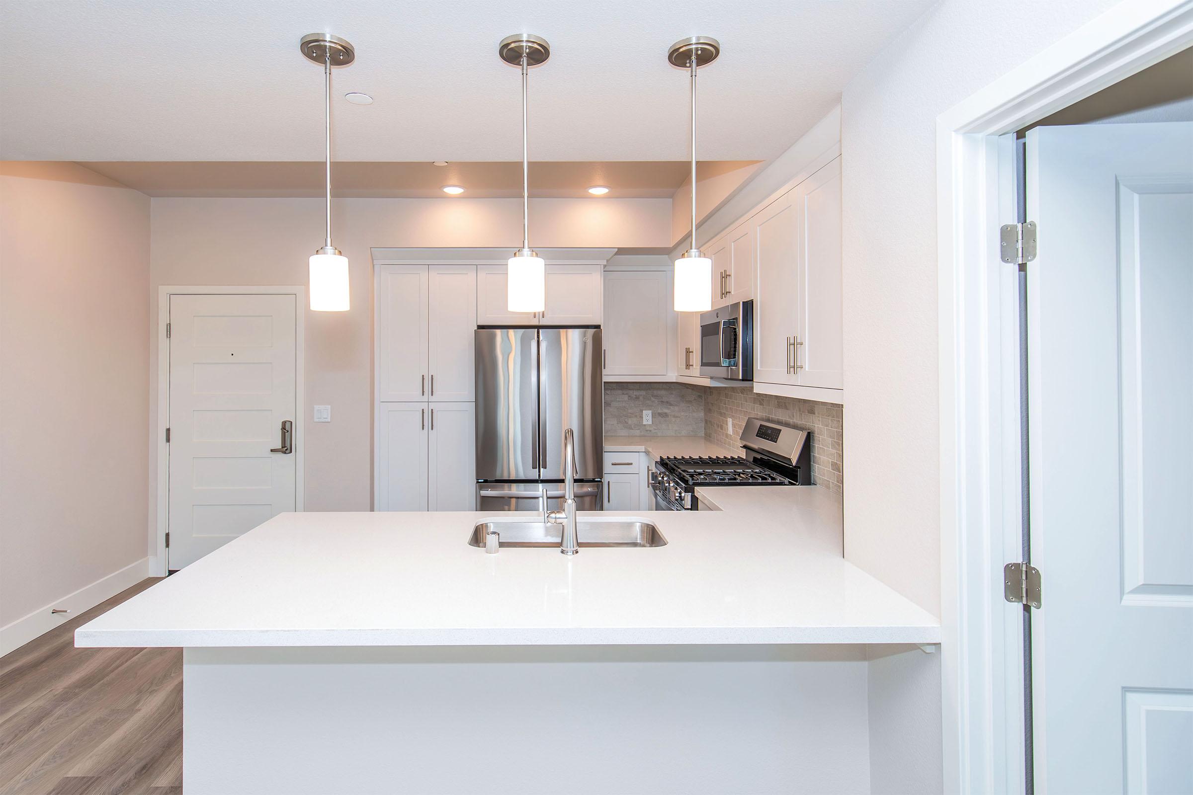 Modern kitchen featuring sleek white cabinetry, stainless steel appliances, and a large island with a sink. Three pendant lights hang above the island, illuminating the space. The flooring is a light wood, and the walls are painted in a soft neutral color, creating a bright and airy atmosphere.