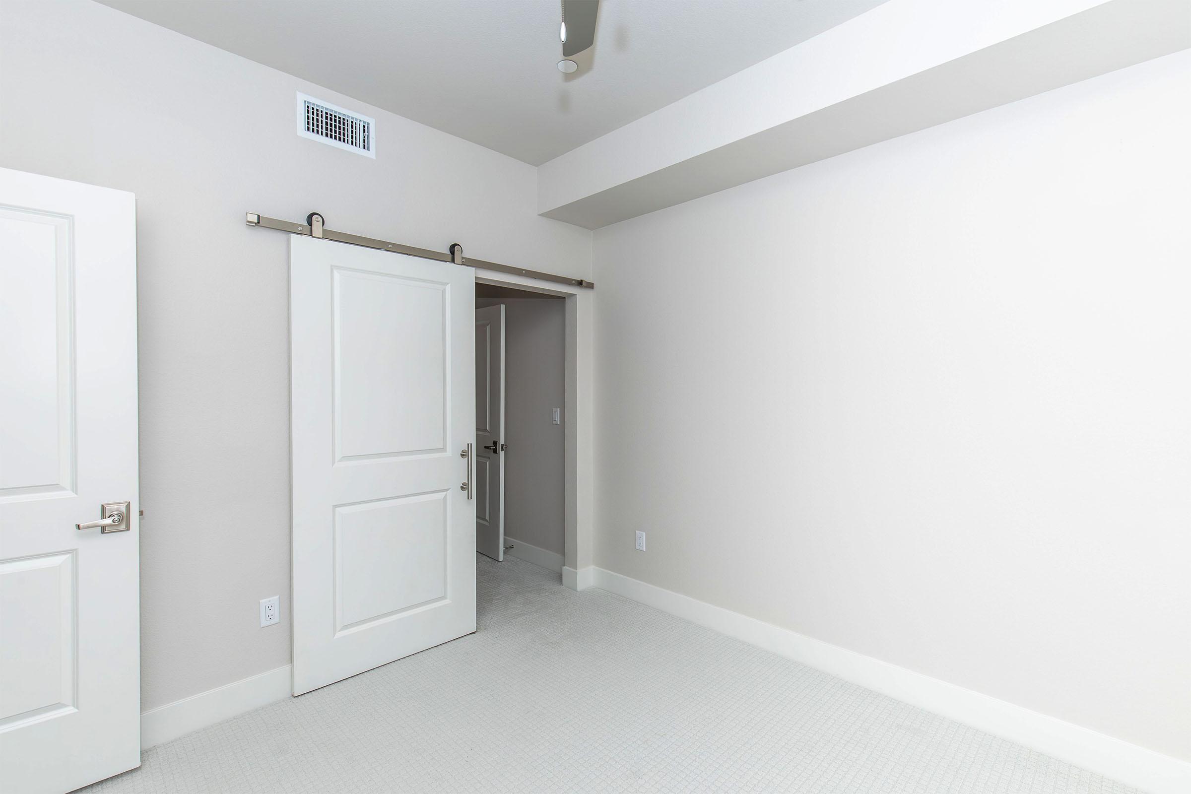 A minimalist room with light gray walls and carpet. A white sliding door is partially open, leading to a small adjoining space. The ceiling features a modern fan, and the room is well-lit with natural light. There are no furnishings present, creating a clean and open feel.