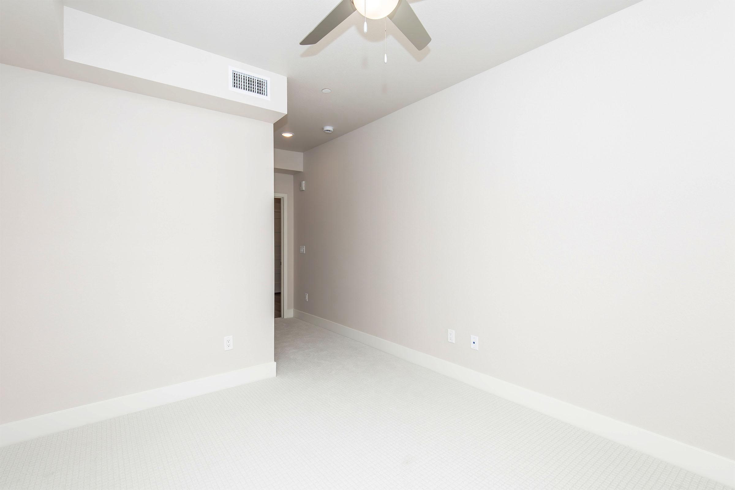 A minimalist room with light-colored walls and carpet. The space includes a ceiling fan and a doorway leading to another area. The overall aesthetic is clean and modern, with no furniture or decorations present, creating an open and airy feel.