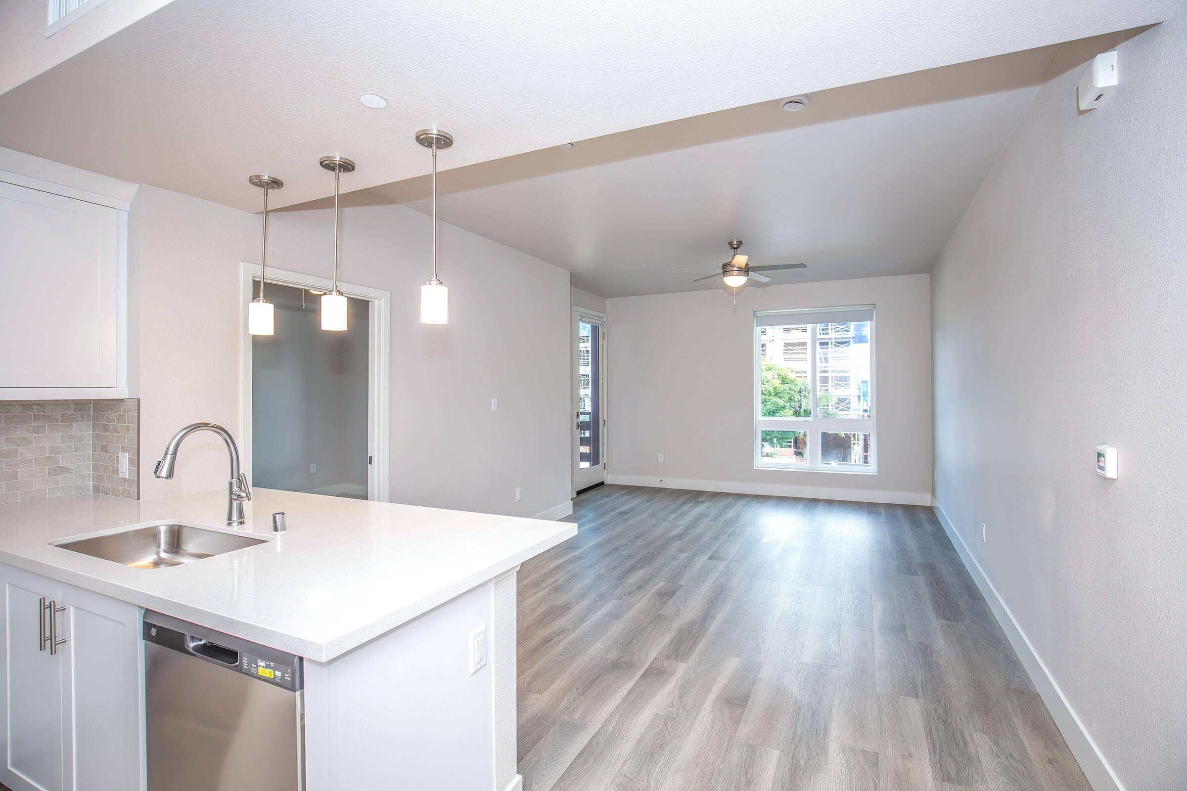 Bright and modern open-concept living space featuring a kitchen with stainless steel appliances and a breakfast bar. Large windows provide natural light, and the room includes light hardwood flooring and a ceiling fan. The area is freshly painted in neutral tones for a contemporary feel.
