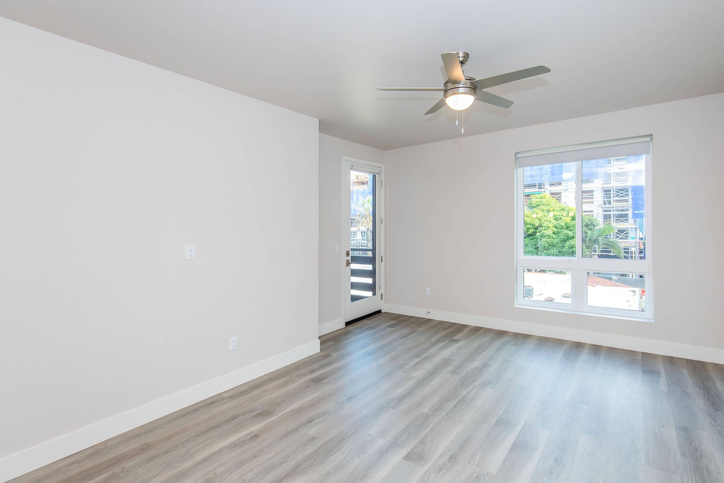 A bright, unfurnished room featuring light-colored walls, large windows, and a ceiling fan. There is a door leading to an outdoor area, and the floor is covered with light-colored wood-like laminate. The space is minimal and modern, ideal for personal decoration.
