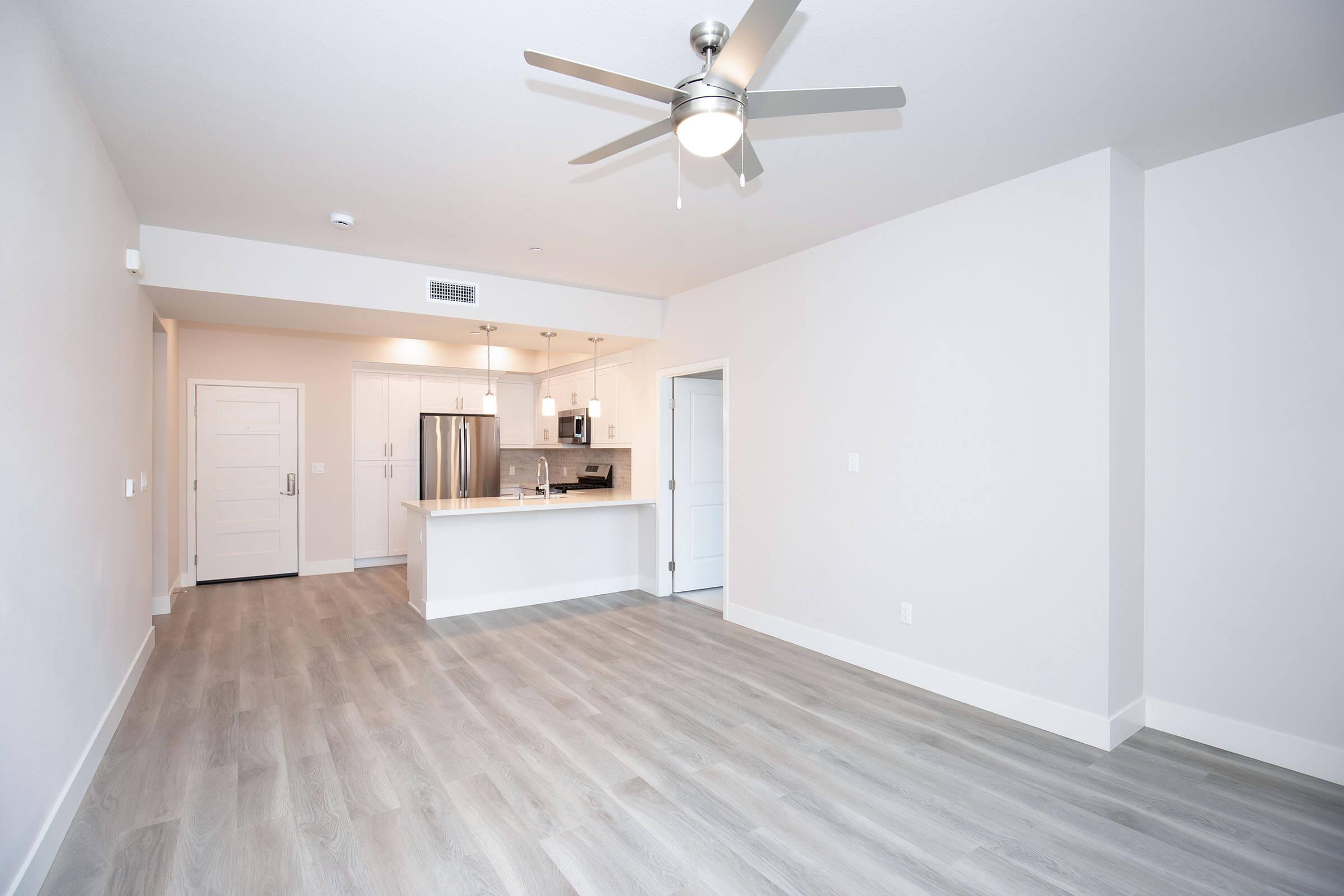 A spacious and modern living area featuring light-colored walls, a ceiling fan, and gray flooring. To the left, there is an open kitchen with stainless steel appliances and a white countertop. The entrance door is visible in the background, leading to a bright hallway.