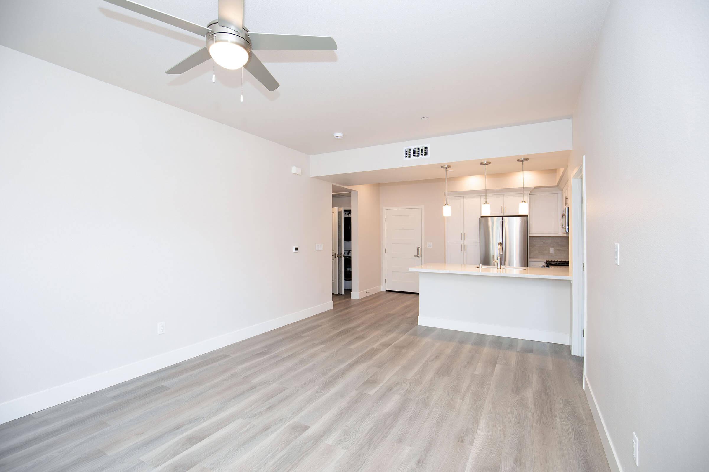 A modern, open-concept living space featuring light-colored walls, light wood flooring, and a ceiling fan. The kitchen is partially visible with white cabinetry and stainless steel appliances. The room is illuminated with natural light, creating a bright and airy atmosphere.