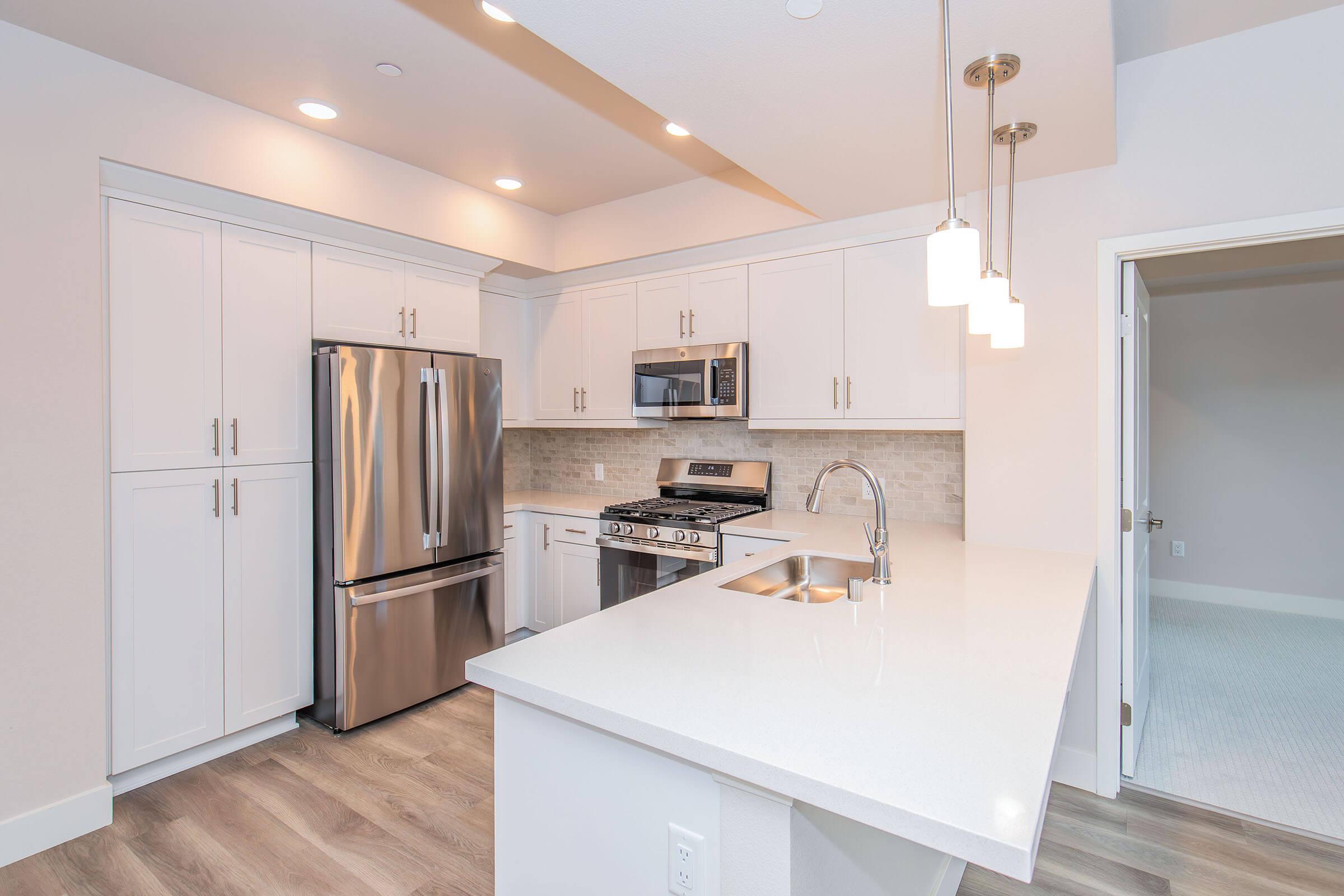 Modern kitchen featuring white cabinetry, stainless steel appliances including a refrigerator and gas stove, a microwave, and a large stone countertop. Pendant lights hang above the kitchen island, and the space is well-lit with neutral tones and a sleek design. Doorway leads to another room.