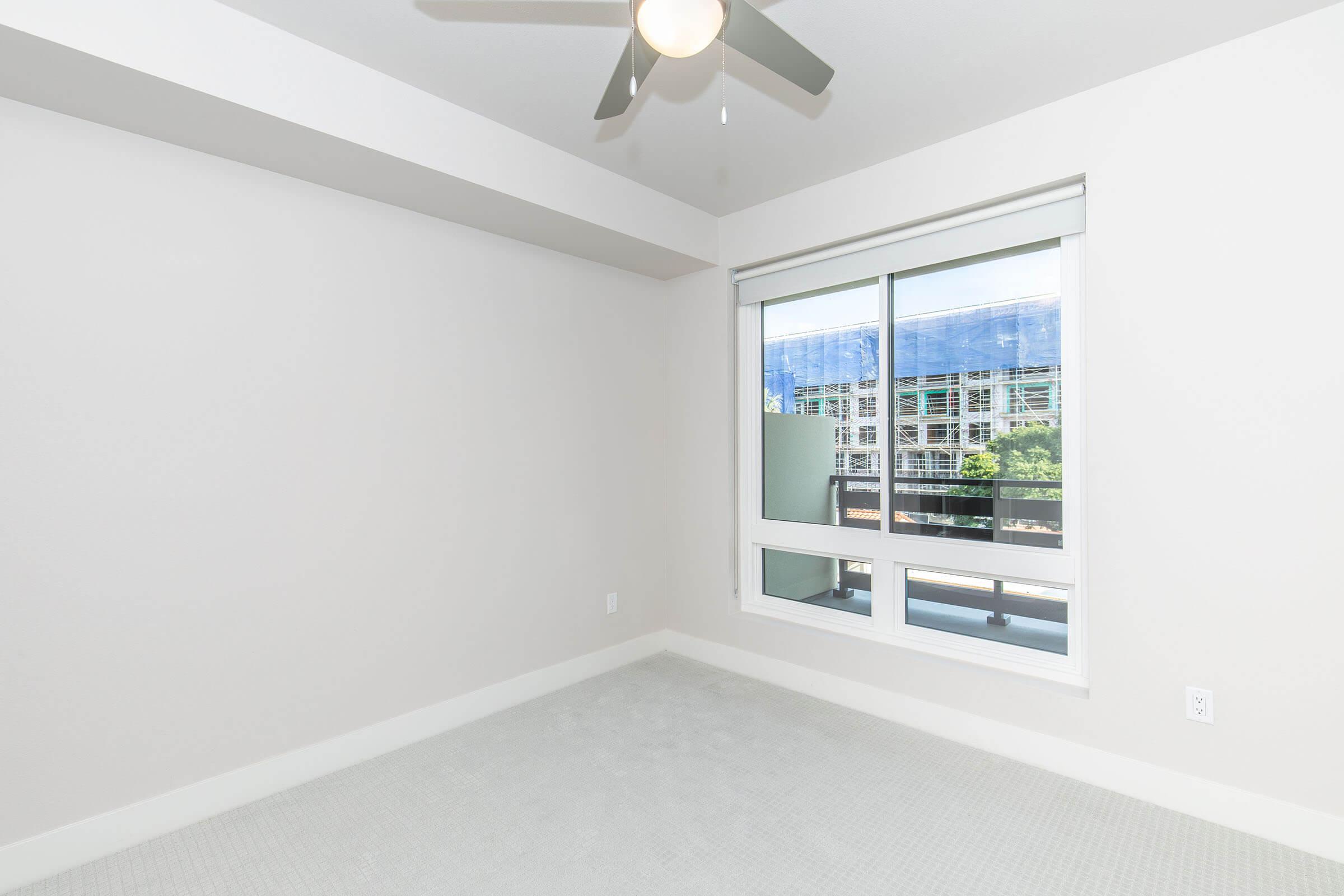 A bright, empty room with light-colored walls and a ceiling fan. There is a large window showcasing views of a building exterior and greenery outside. The floor is covered with a light, carpet-like material. The overall atmosphere is clean and minimalistic.