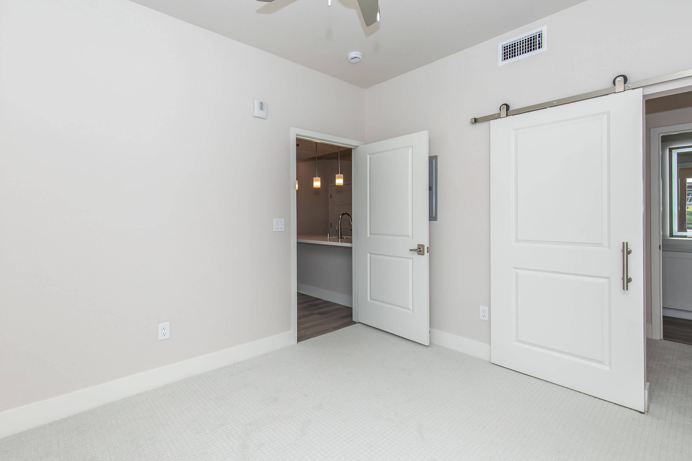 A spacious, well-lit room with light-colored walls and carpet. Two white doors, one partially open, lead to another area. The room features a ceiling fan and a neutral aesthetic, creating a clean and modern look.