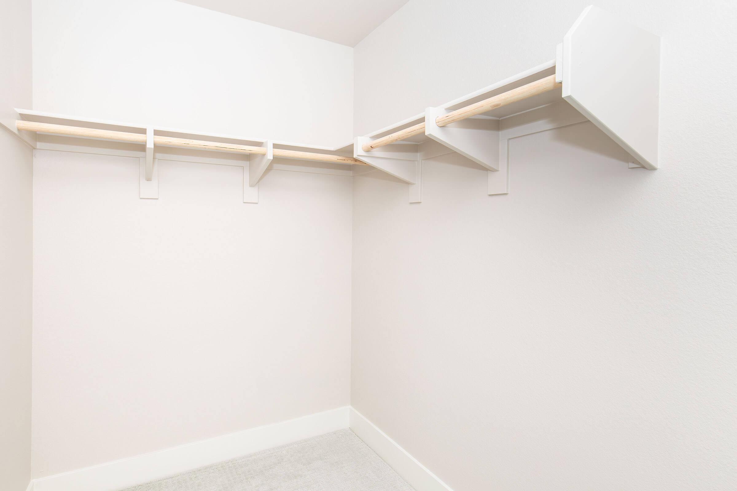 An empty closet featuring white shelving with wooden support beams, set against a light-colored wall. The design is minimalist, with clean lines and ample space for storage. A soft carpet covers the floor, enhancing the room's simplicity and openness.